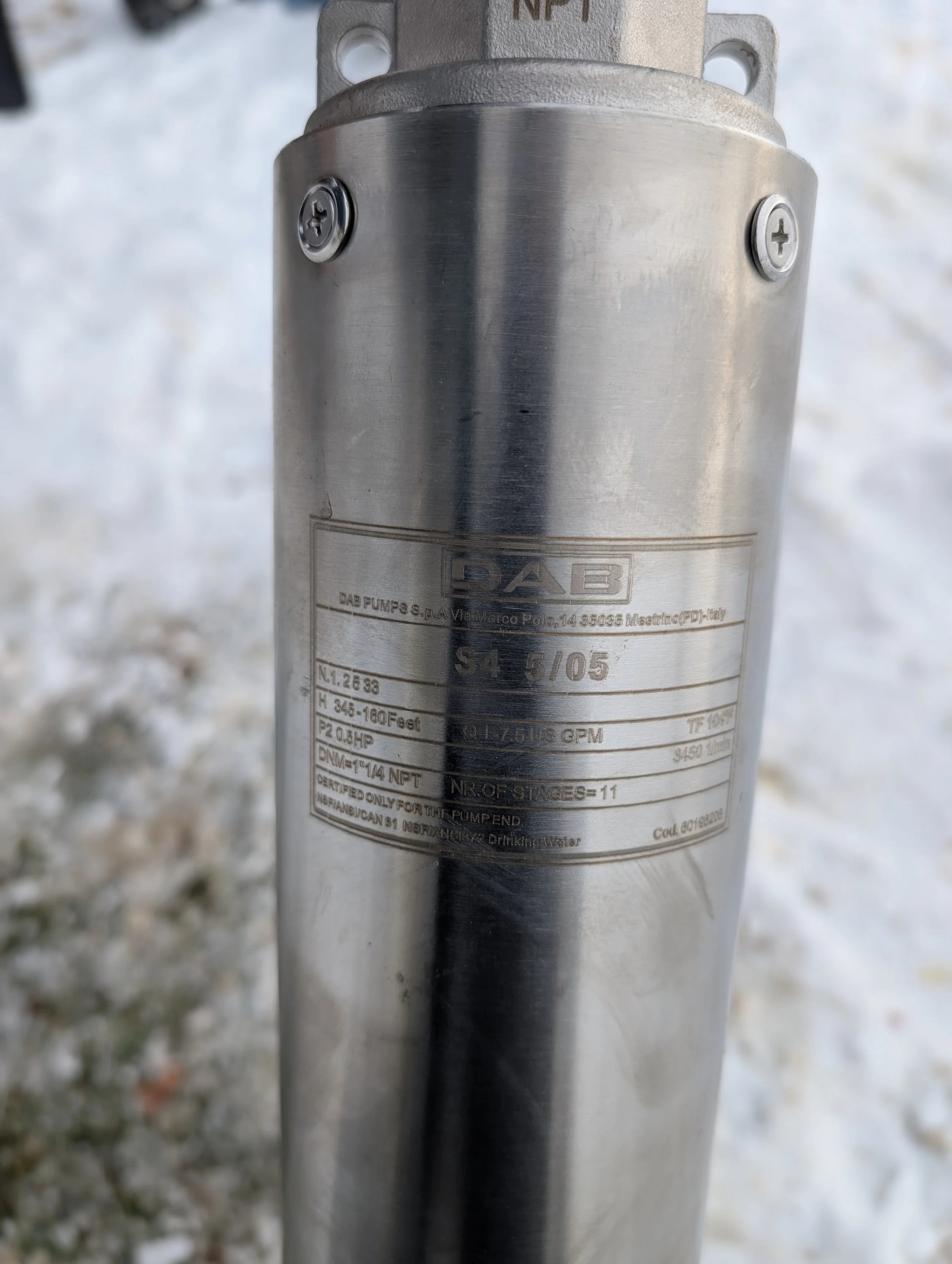 Close-up of a cylindrical metal (probably stainless steel) pump or mechanical device with labels and screws, with a blurred outdoor background.