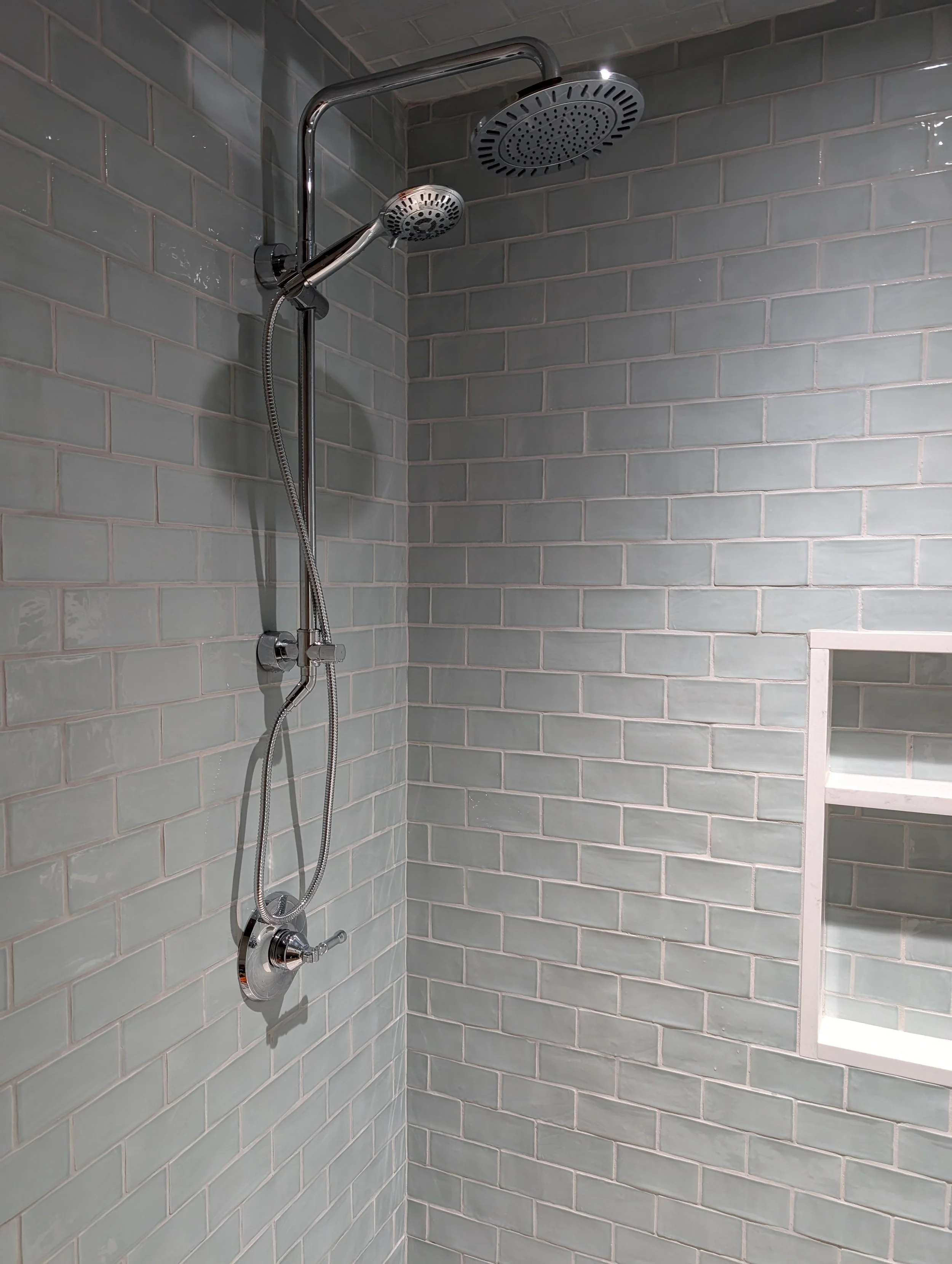 Shower with gray subway tiles wall, silver multi-head shower system, including a large overhead shower head and a detachable handheld shower, in a modern bathroom.