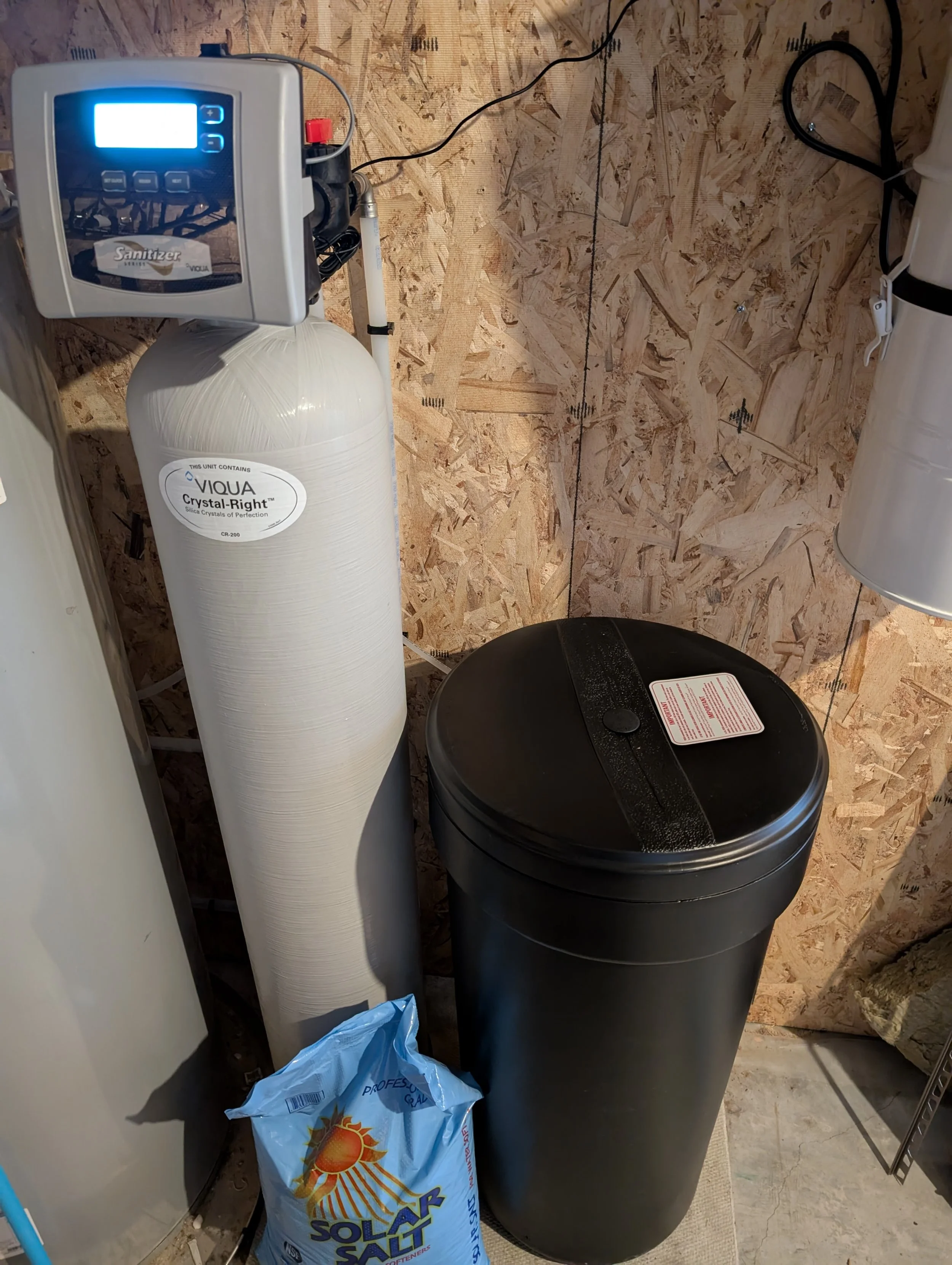 A water softener system with a control valve, salt tank labeled 'VIQUA Crystal-Right,' and a plastic bag of solar salt in a utility room with unfinished wooden wall panels.