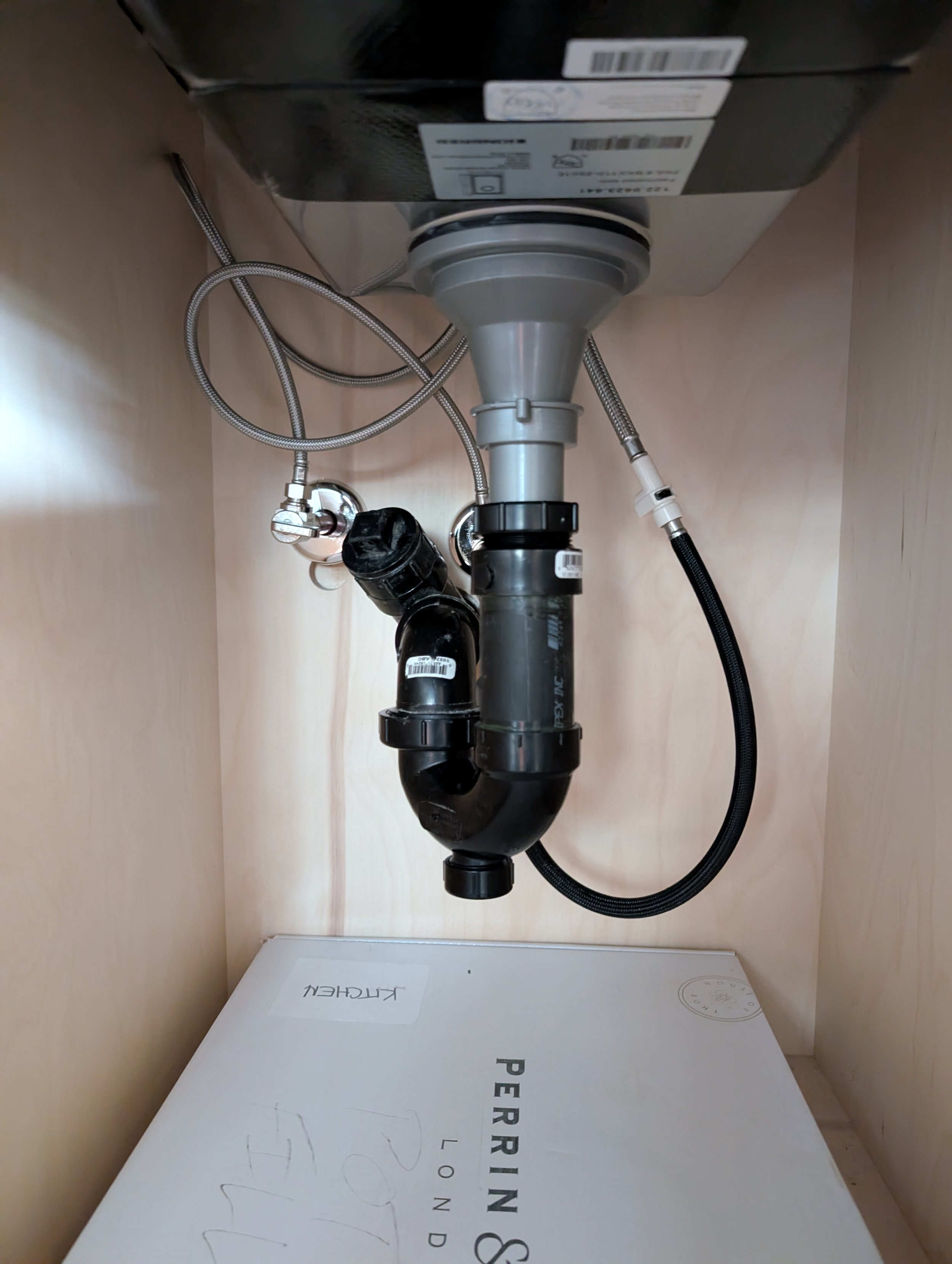 The image shows the underside of a kitchen sink with plumbing pipes, hoses, and a drain assembly. There is a cardboard box labeled "PERFECTING" resting on the floor under the sink. The photo is taken from a low angle.