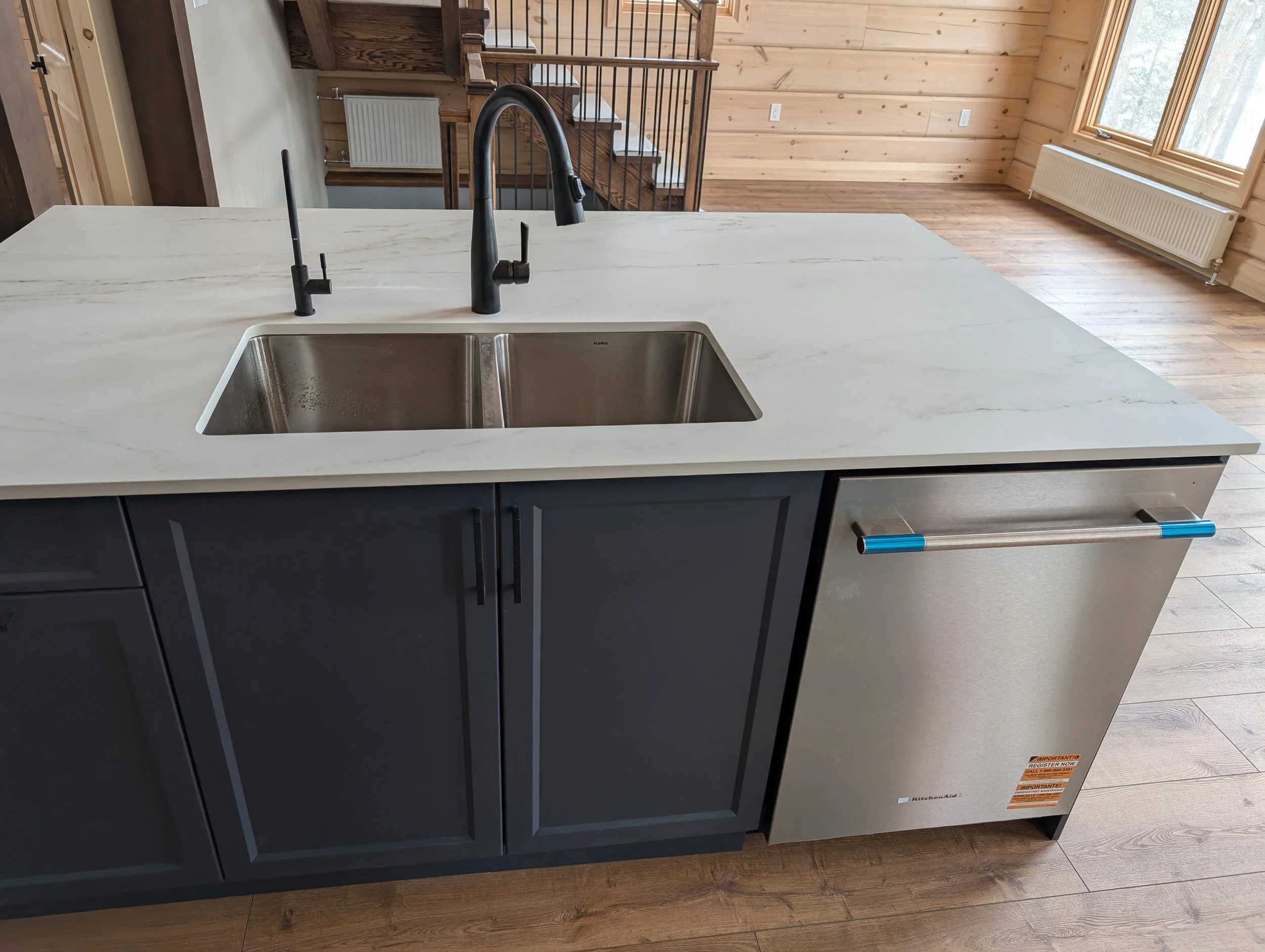 A modern kitchen island with a white marble countertop, stainless steel sink, and matte black faucet and handles in a room with wooden walls and flooring, a staircase, and large windows.