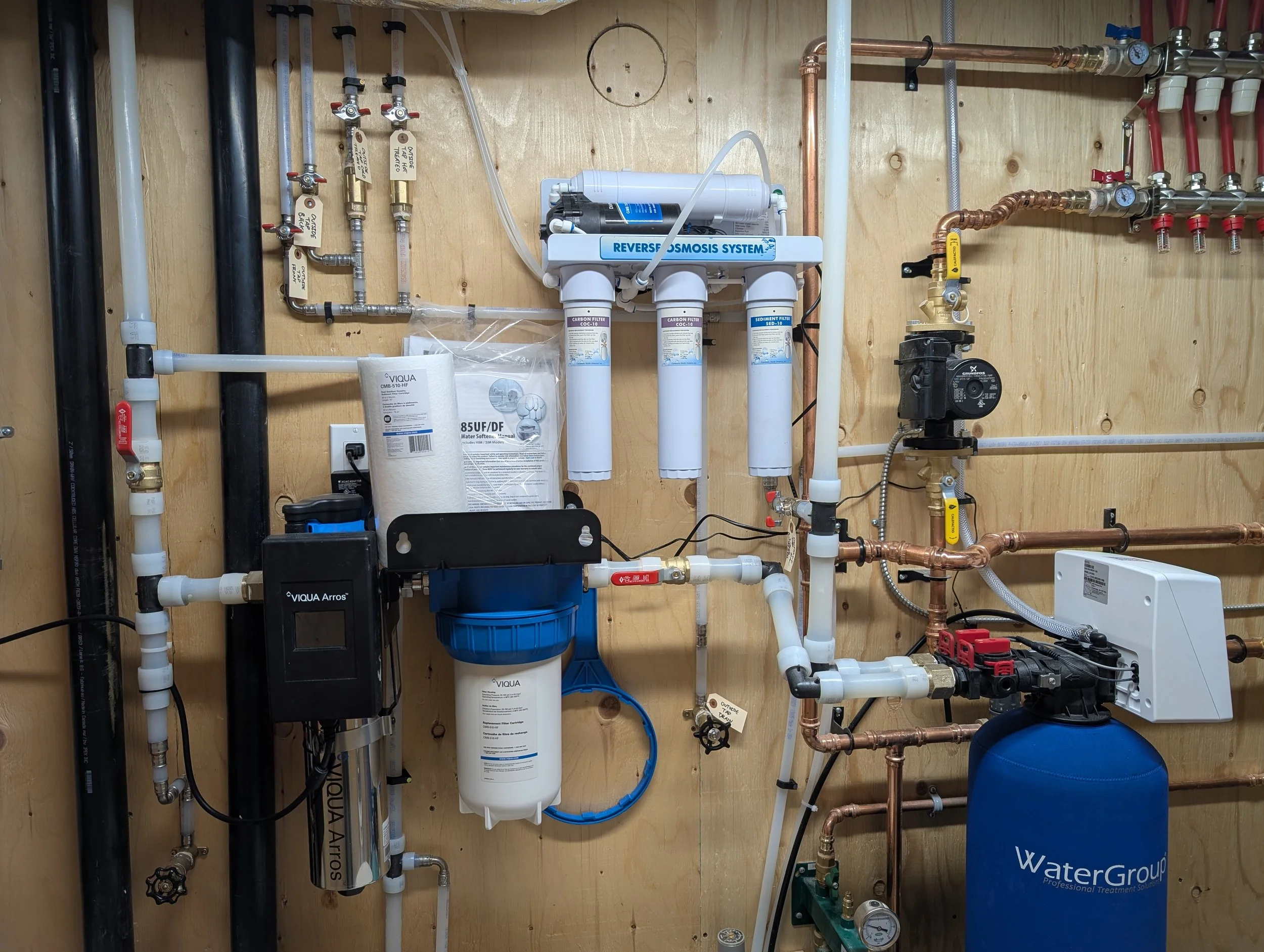 A water filtration system mounted on a wooden wall, comprising filters, pipes, and gauges.
