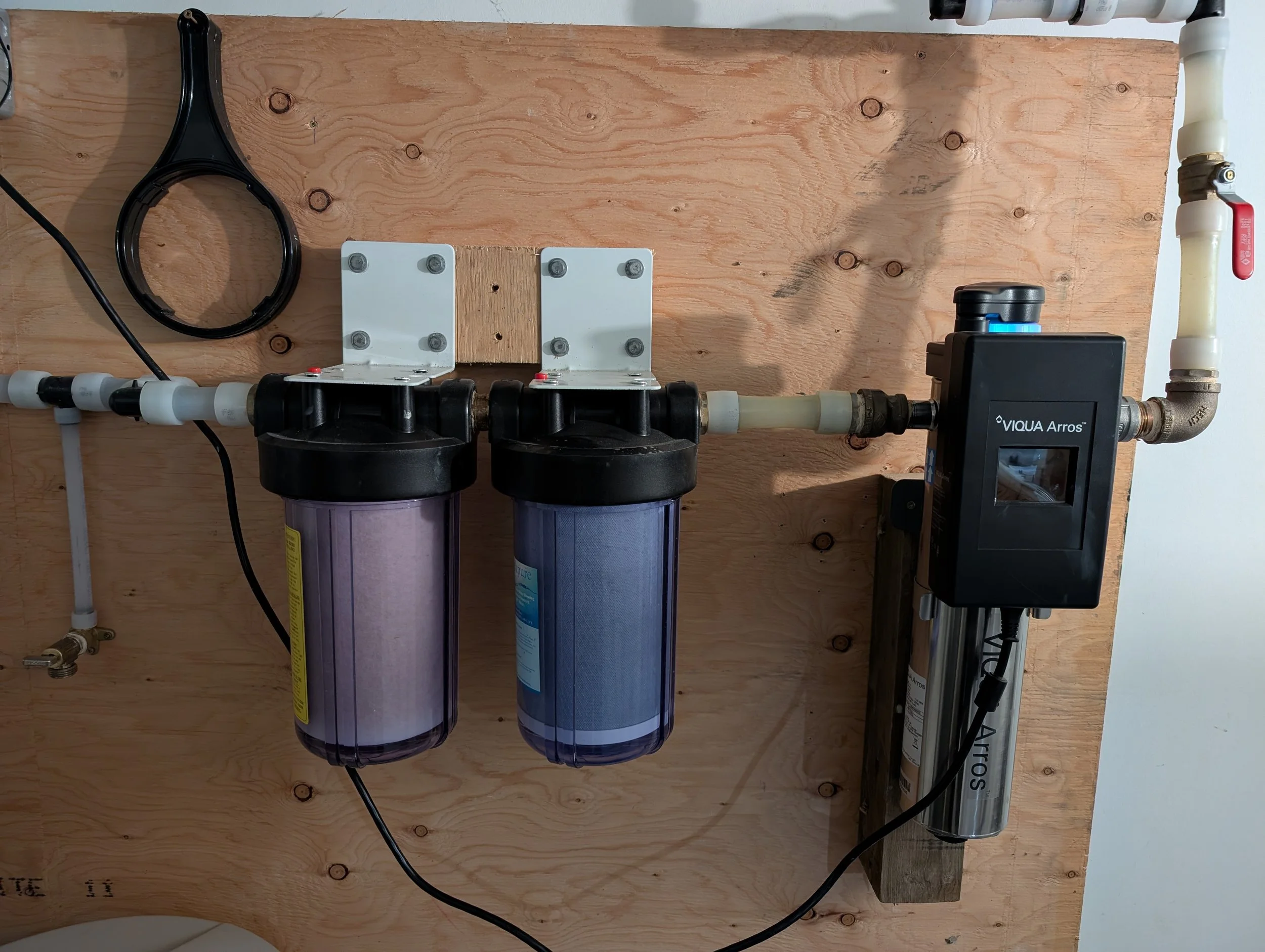 A water filtration system mounted on a wooden wall, consisting of two purple filter canisters, white piping, a black control valve, and a black and blue digital controller labeled 'VIQUA Arros'.