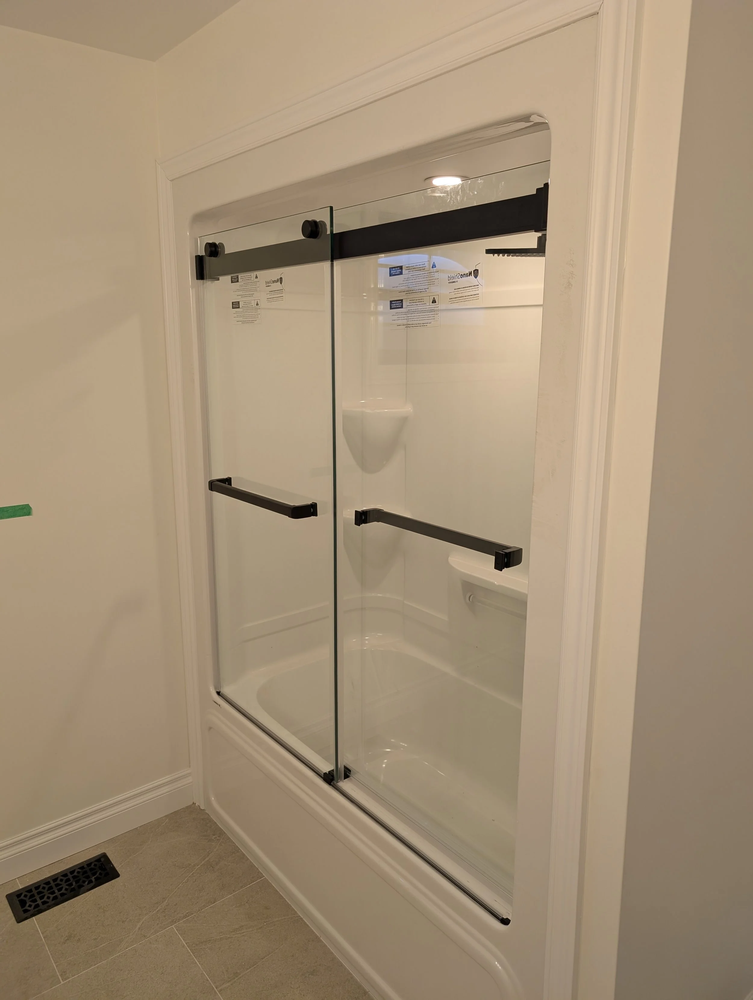 Bathroom shower with glass doors, white walls, a small wall-mounted white urinal, beige tiled floor, and a black vent on the floor near the corner.