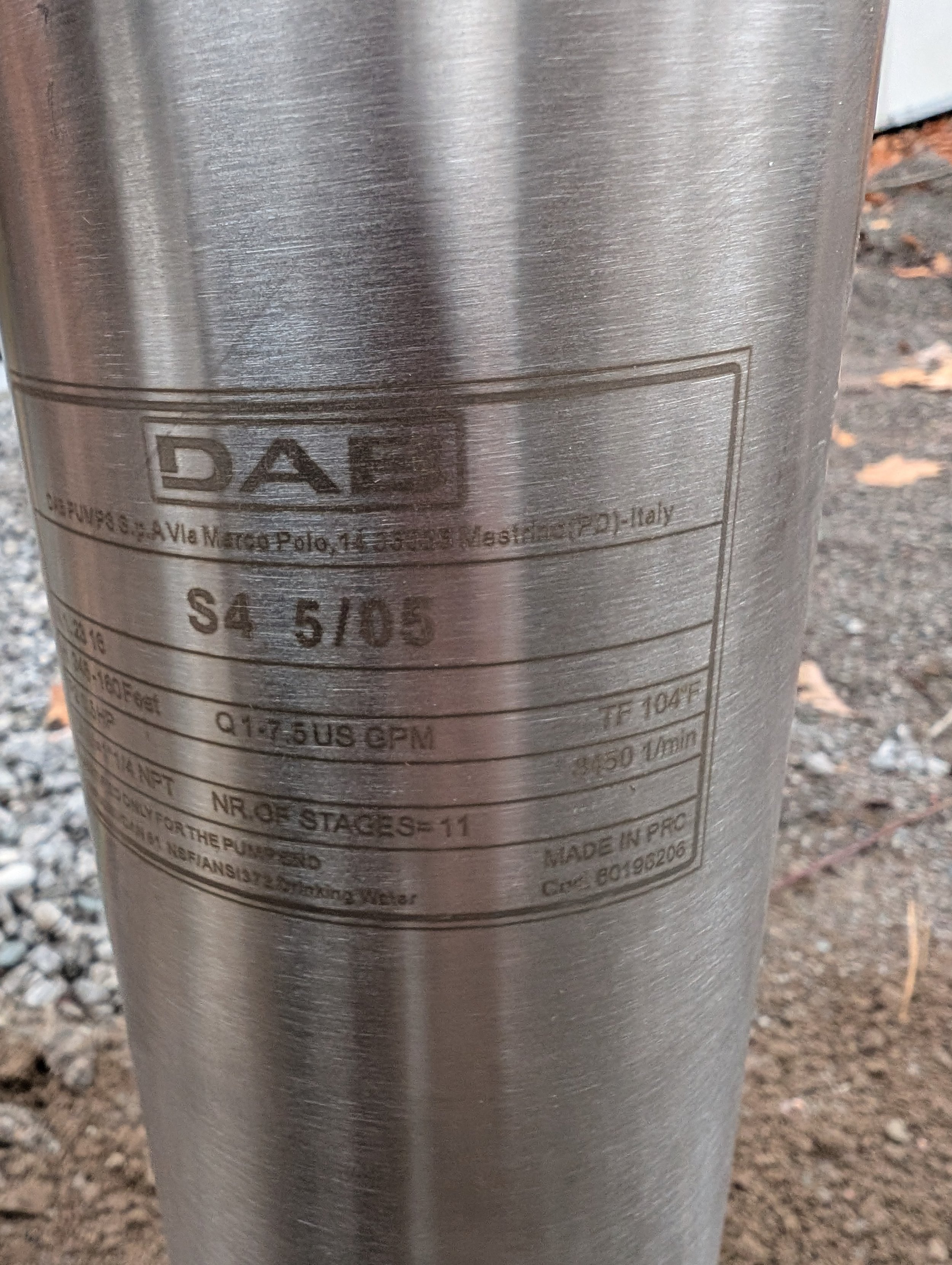Close-up of a metallic cylindrical object with a label that reads 'DAD,' along with other technical and manufacturing information, set outdoors on gravel ground.