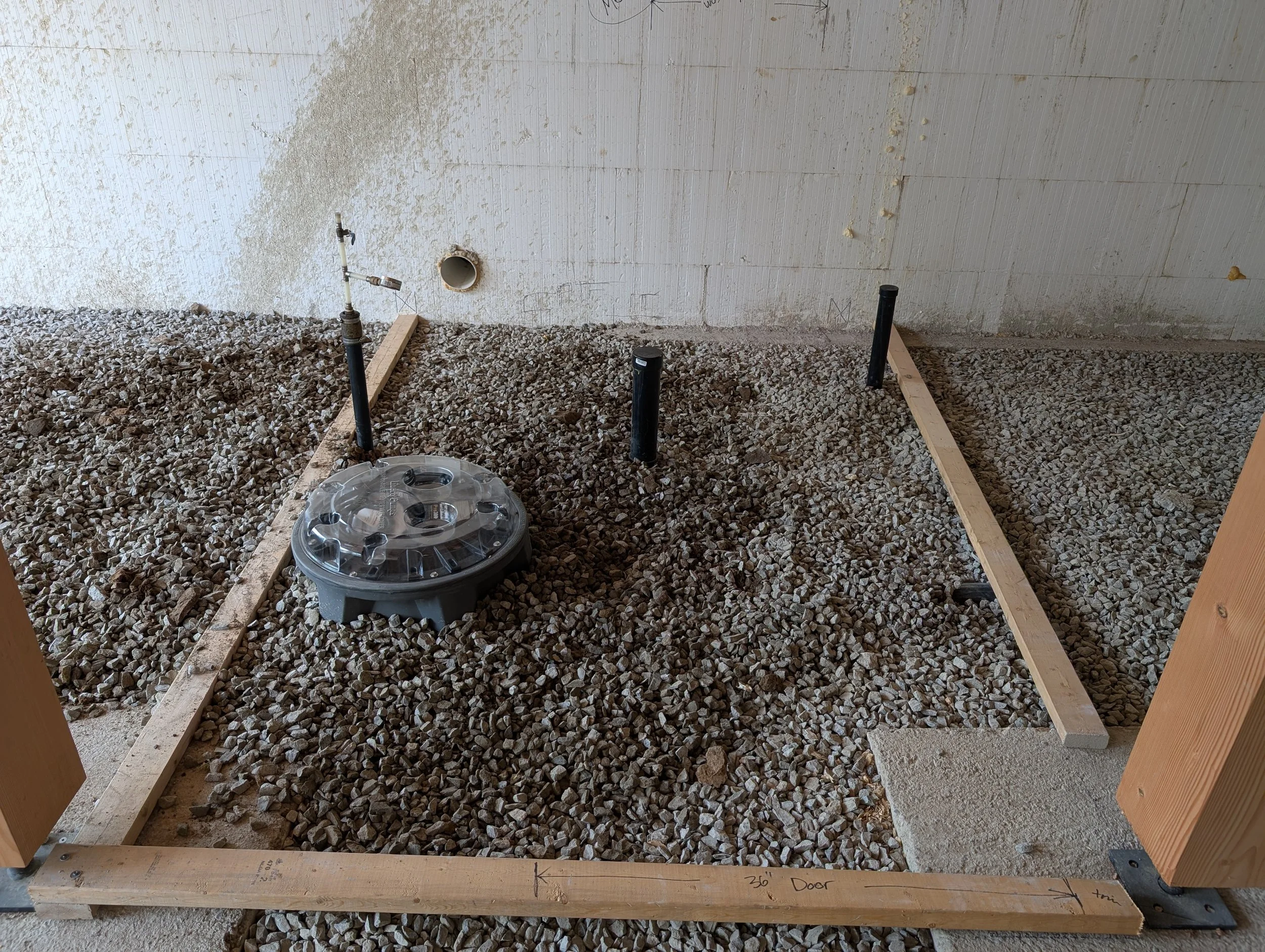 Construction area with gravel floor, black pipes, wooden framing, and a water spigot on a temporary wall.