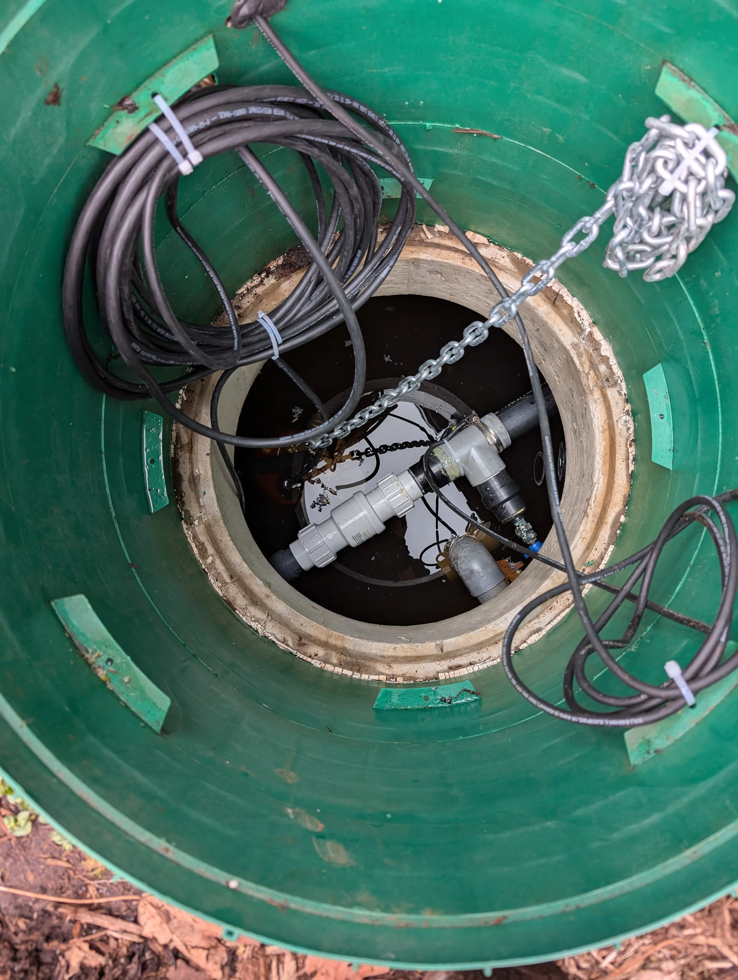 An underground utility or maintenance access point with a green circular cover. Inside, black cables are coiled and secured with cable ties, and a metal chain is attached to the sides. Several pipes and fittings are connected at the bottom, and the c