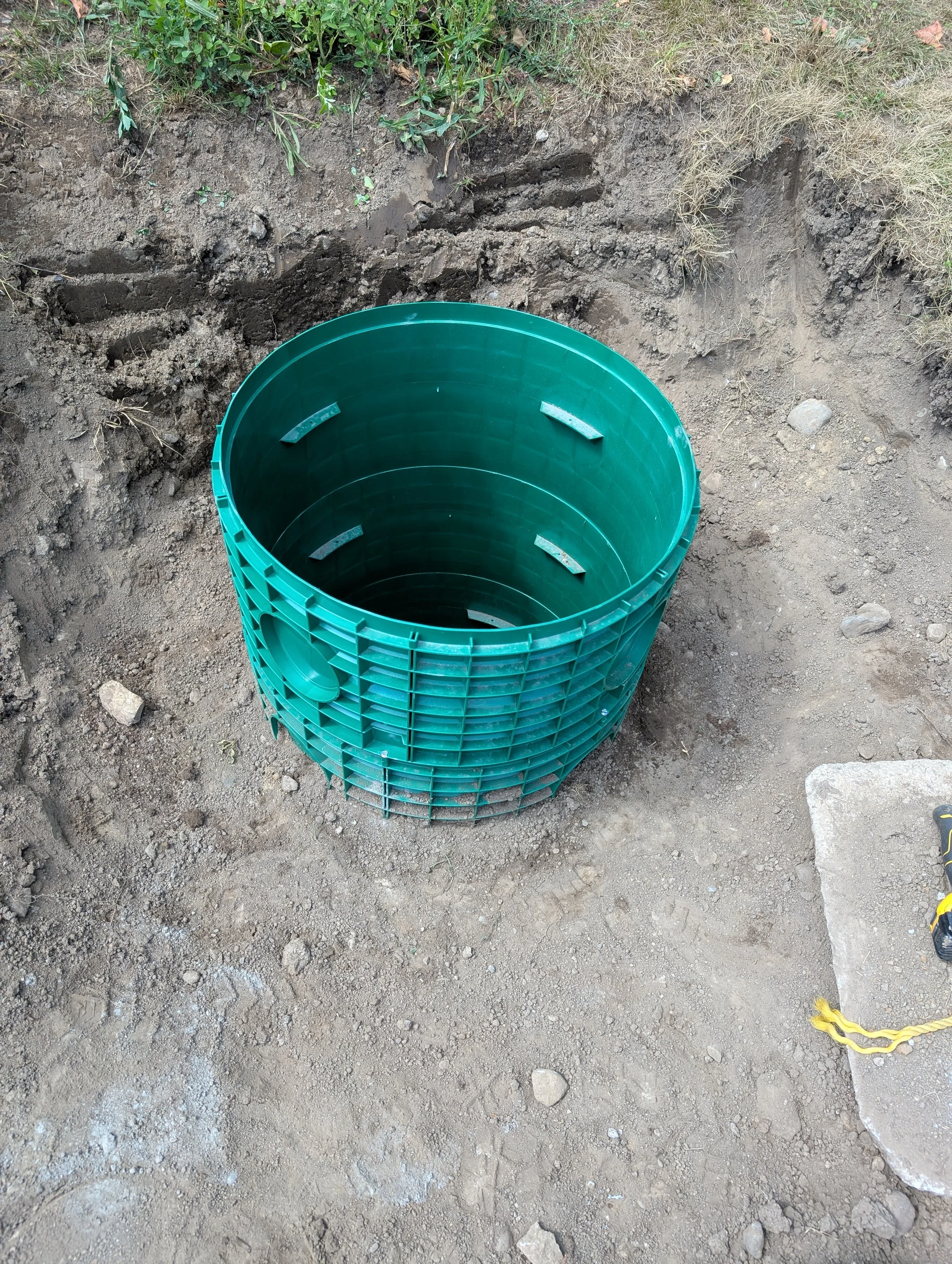 An open green plastic container or bucket situated in a dug-up patch of bare soil and dirt, with patches of grass and small rocks around.