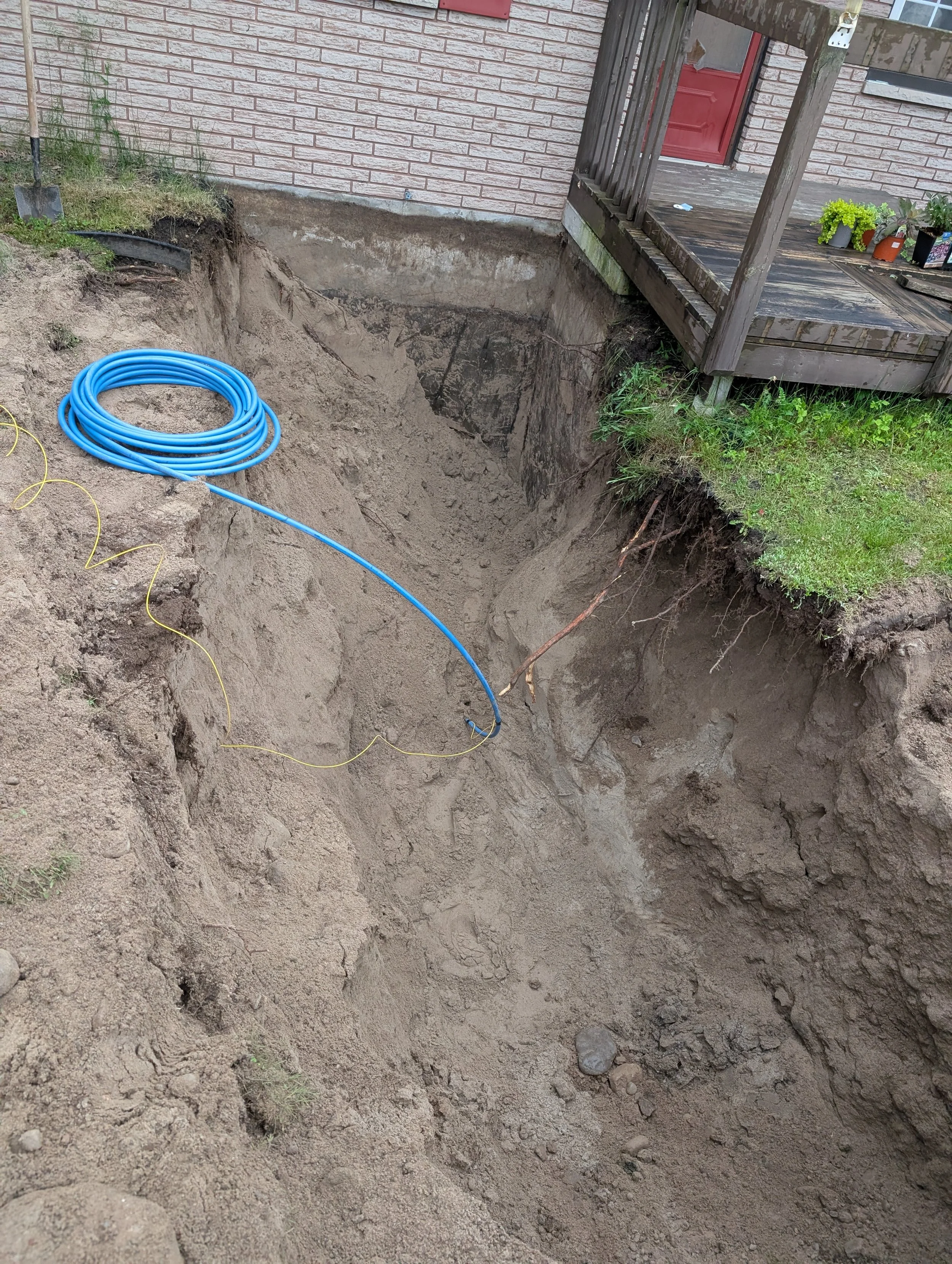 A large hole in the ground next to a house with a brick wall, a small porch with potted plants, and a wooden railing. Construction tools and blue piping are present around the excavation site.