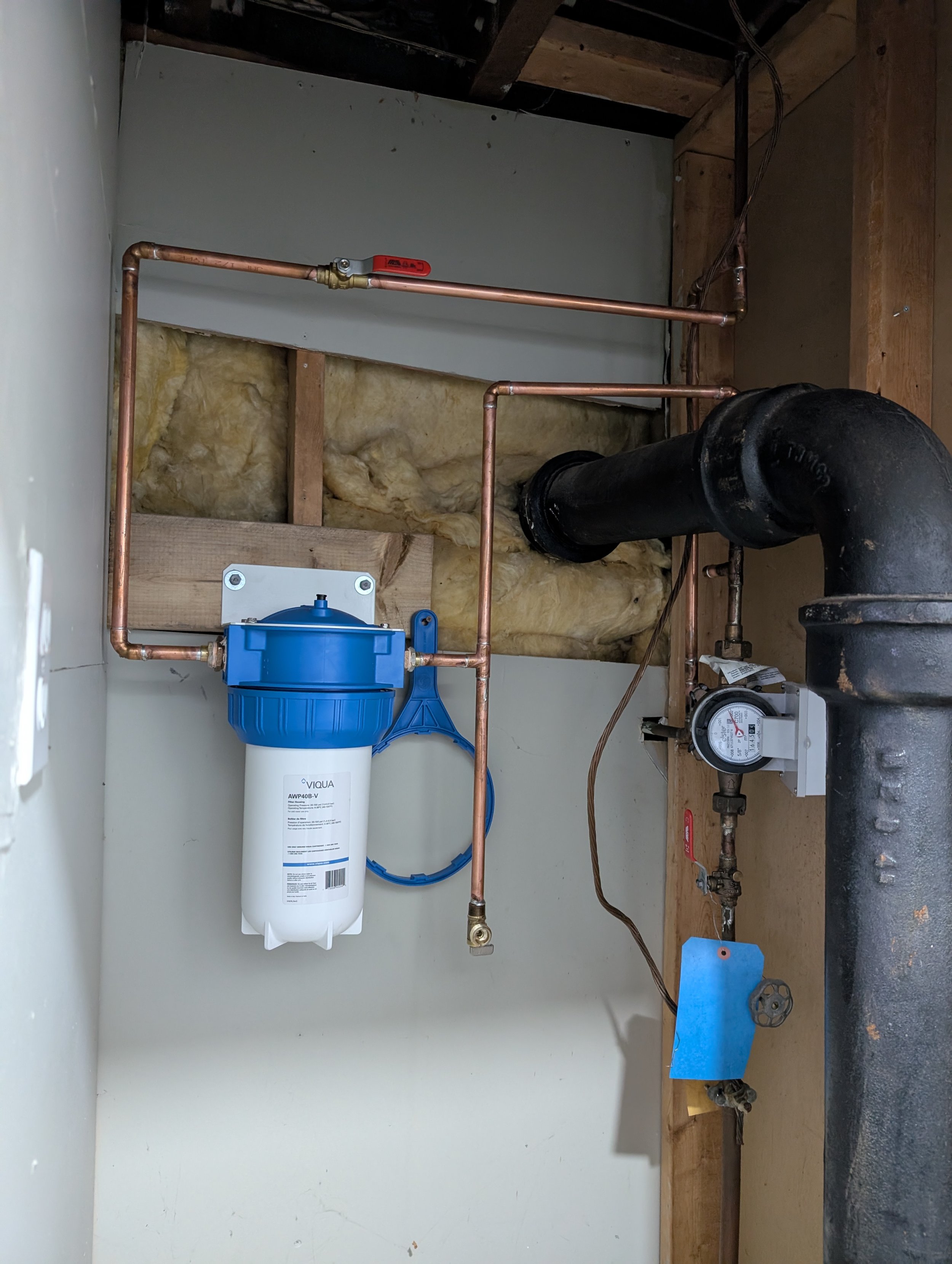 A water filtration system installed in a utility closet with copper piping, an automatic shut-off valve, a black pipe, and an electrical meter on a wooden surface.
