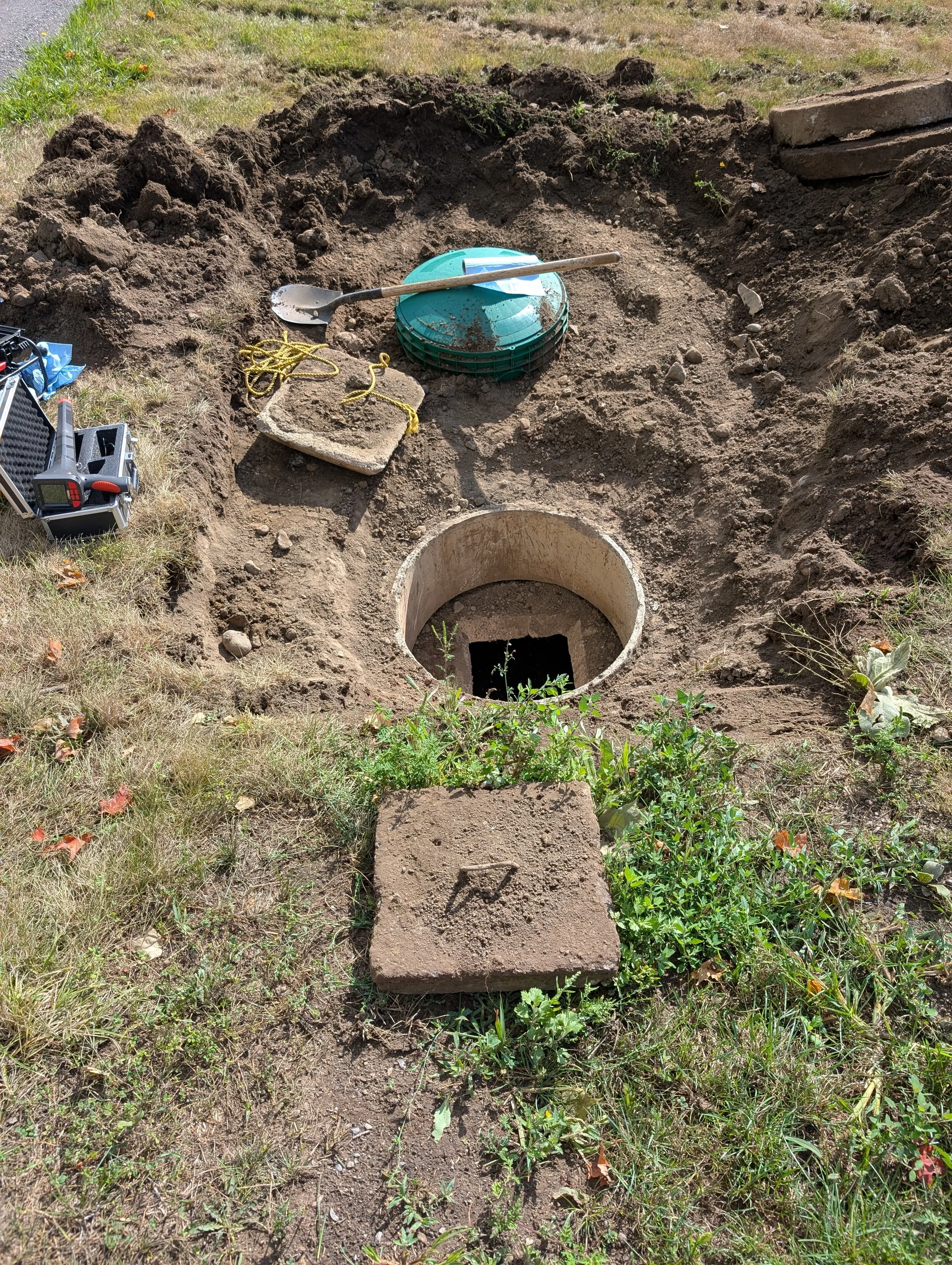 A construction site with a circular hole in the ground, some soil, a shovel resting on a stack of green lids, and a concrete block with a yellow rope on top.