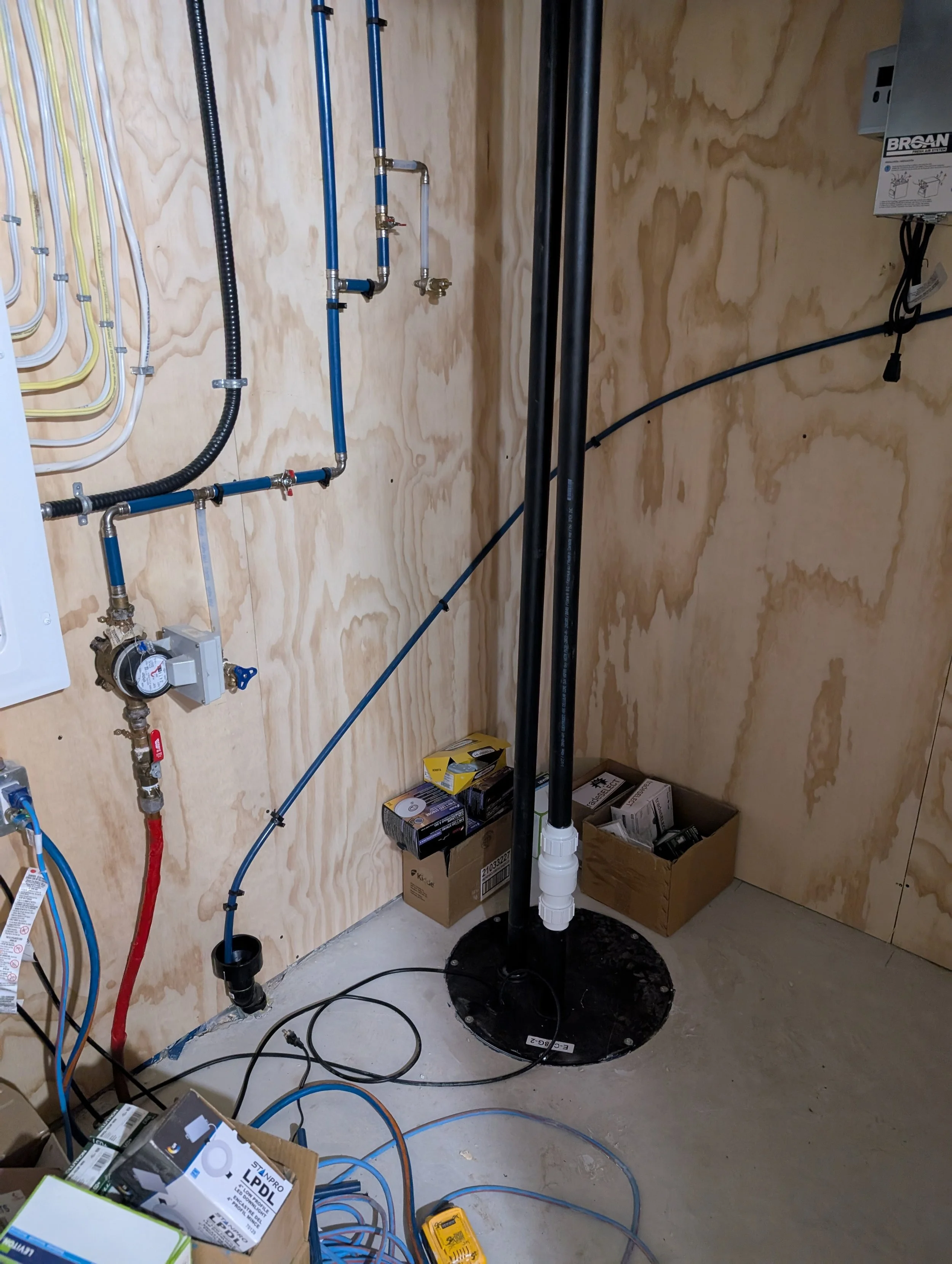 Interior of a utility or laundry room with plumbing, pipes, and electrical wiring being installed; boxes and tools on the floor.