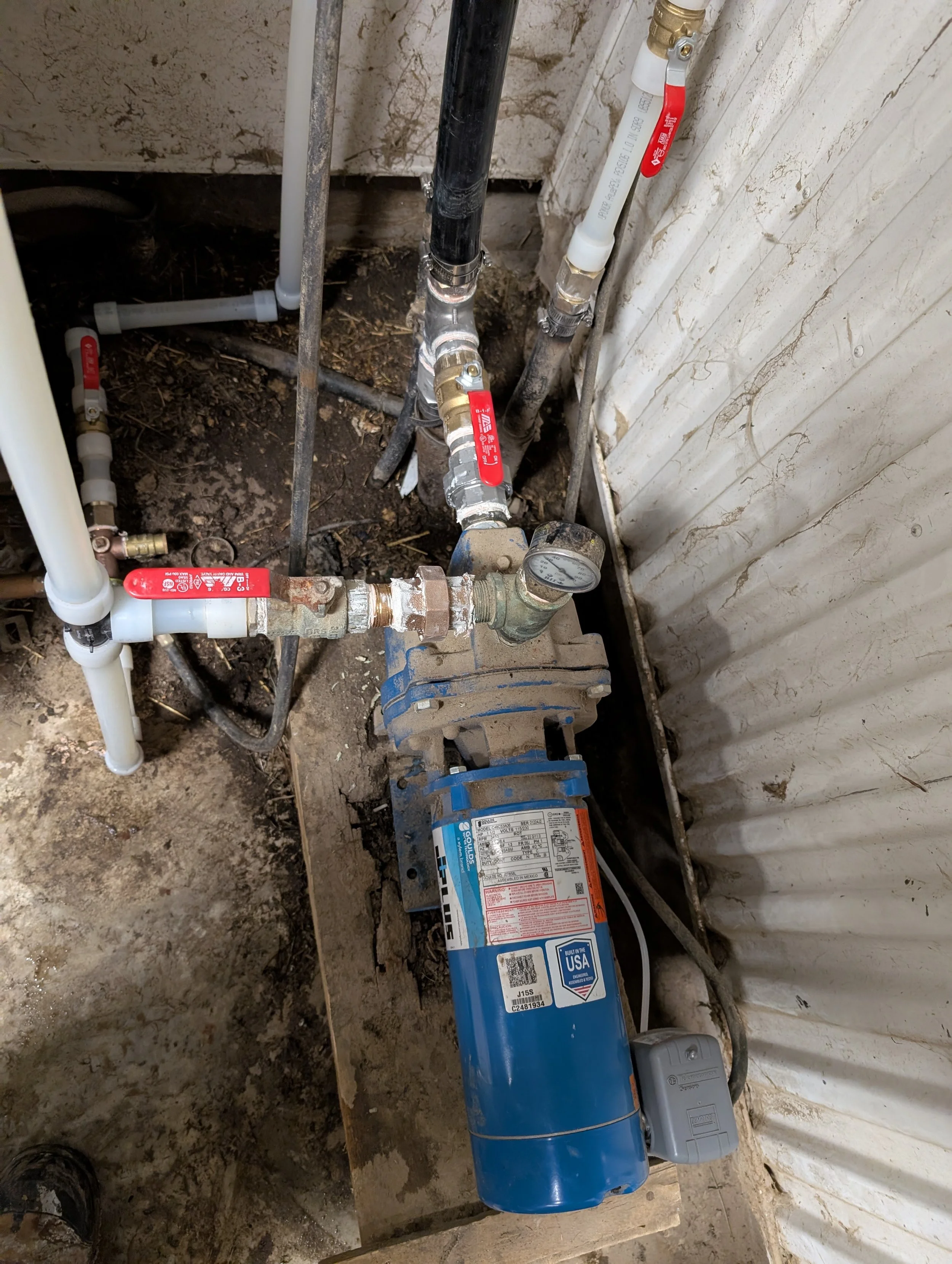 A water pump system with pipes and valves installed in a construction area with dirt floor and unfinished wall.