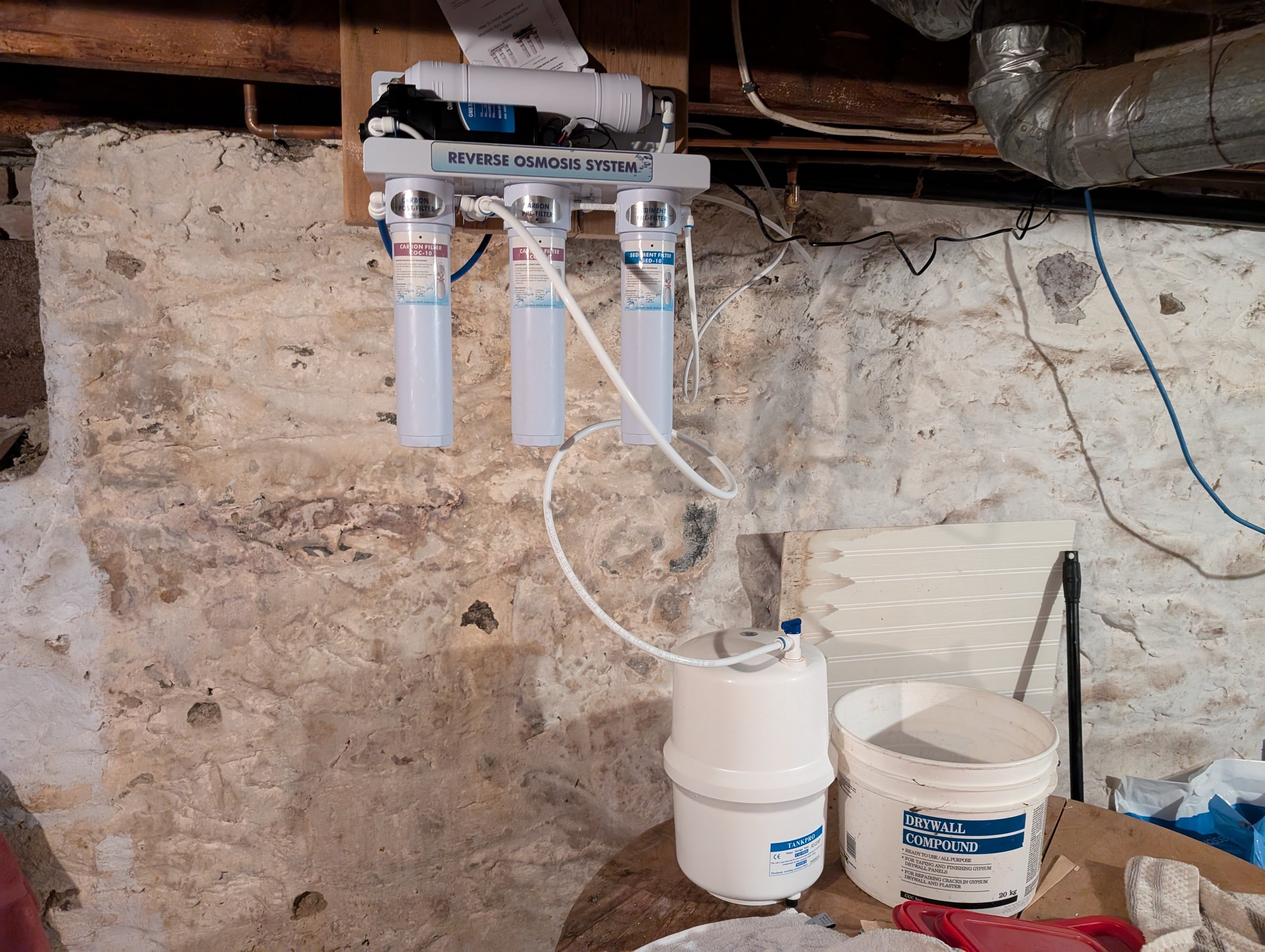 A reverse osmosis water filtration system mounted on a stone wall in a basement or utility room, with filters and pipes connected, and containers of drywall compound and other items nearby.