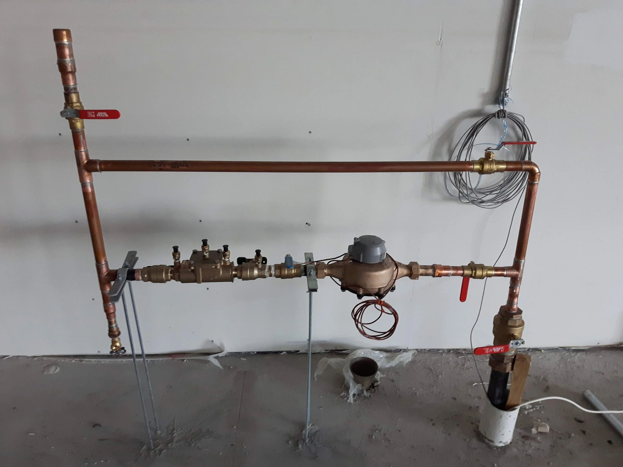 Copper plumbing pipes with shut-off valves on a wall, electrical wiring and a pressure regulator, under construction.