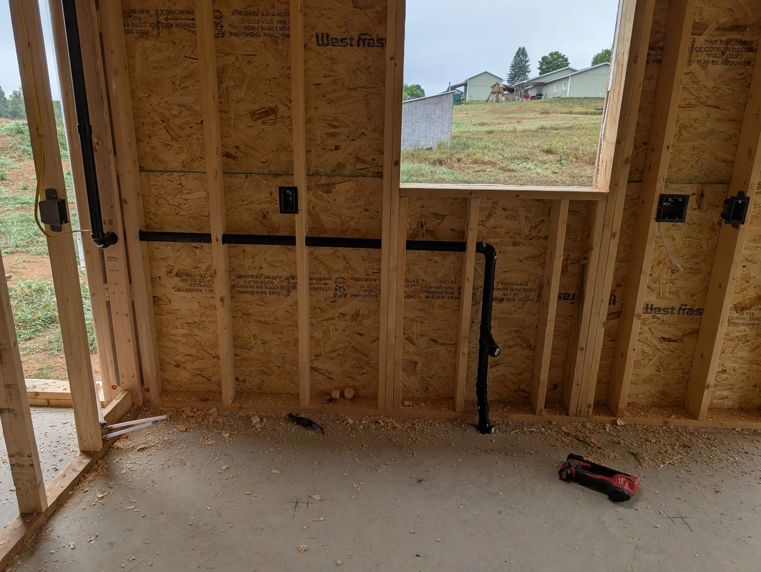 Interior of a framed house with wooden studs, electrical outlets, and pipes, overlooking an outdoor grassy area with neighboring houses.