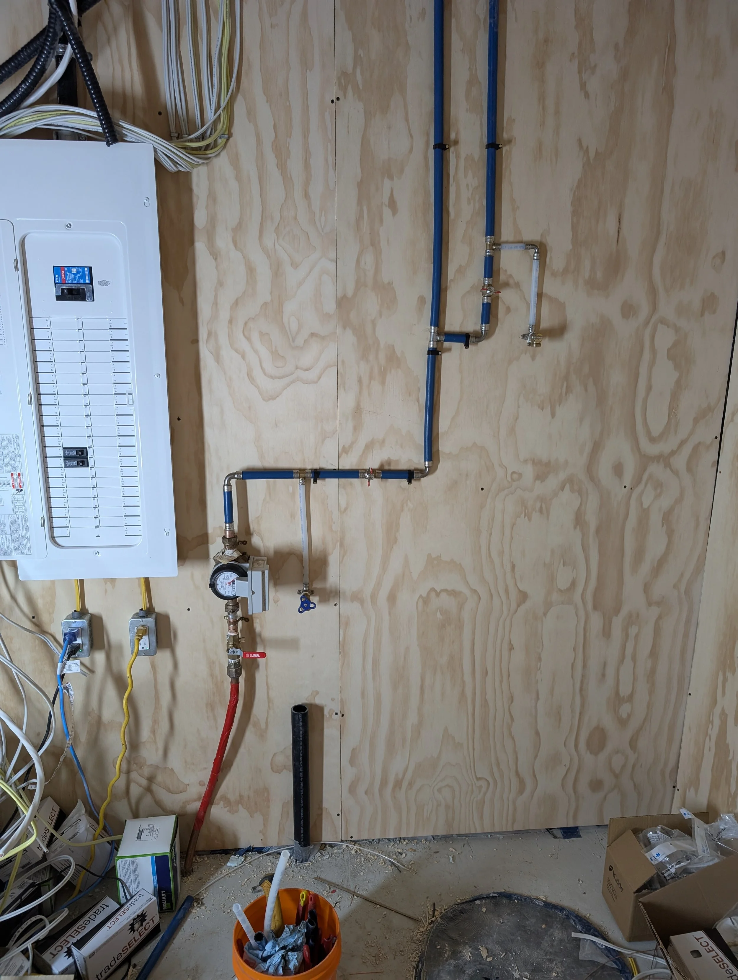 Construction or repair site showing a bathroom wall with plumbing pipes, an electrical panel, wires, and various construction tools and materials.