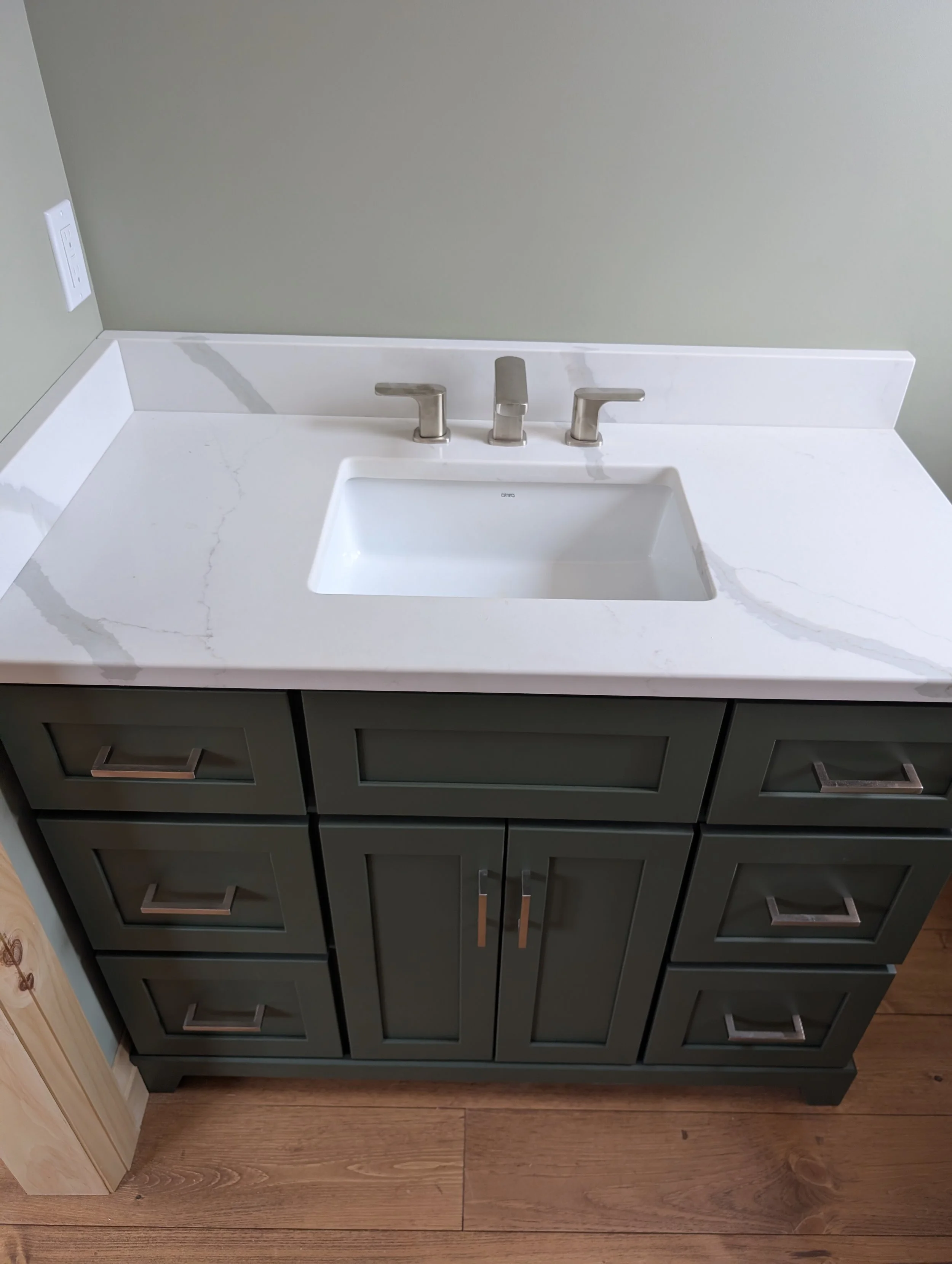 A bathroom vanity with a white marble countertop, an under-mount sink, and a gray cabinet with silver handles. There is a wall outlet on the left side.