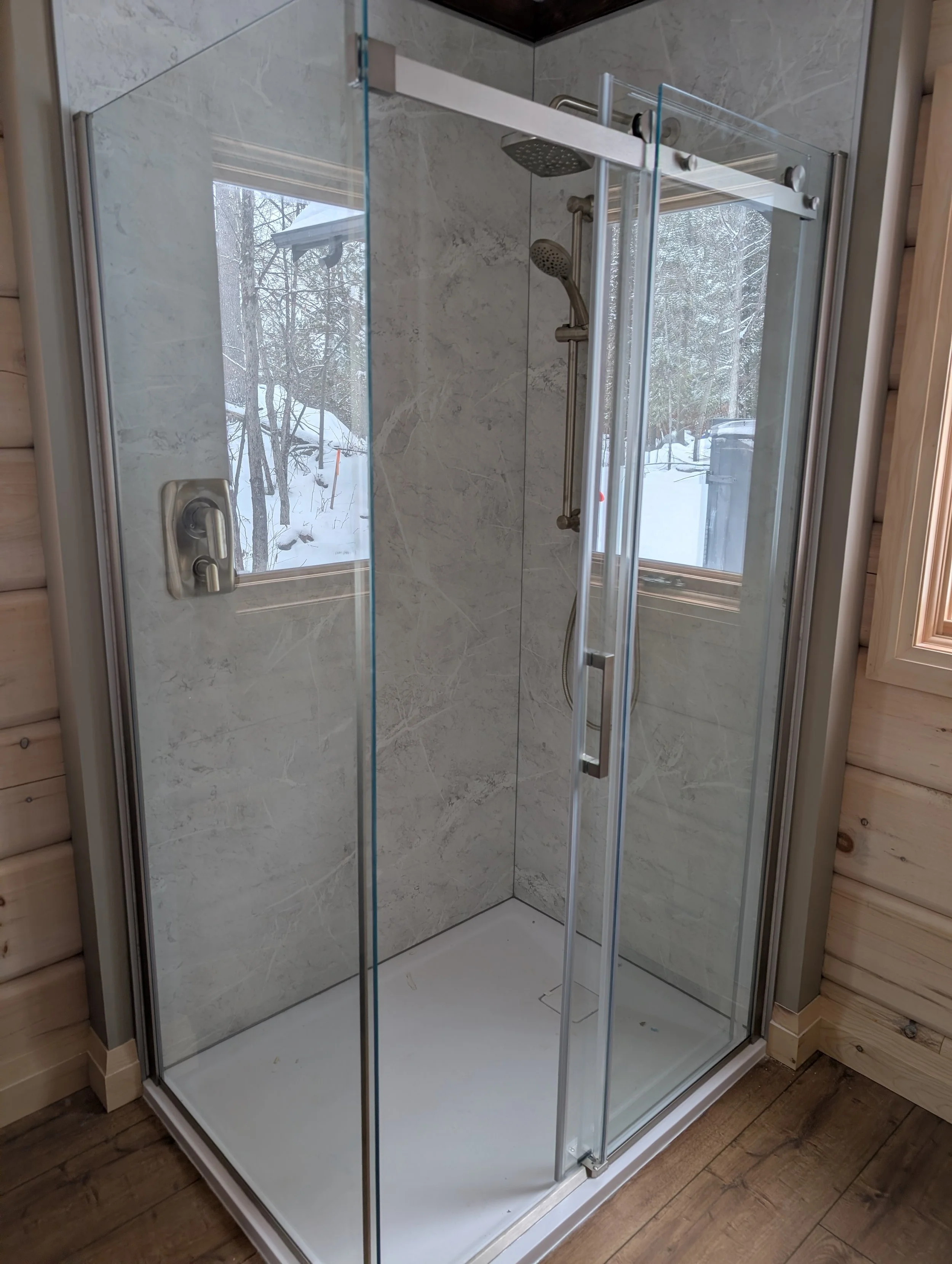 A glass shower stall with marble wall tiles, a rain shower head, and a handheld shower head. It is situated next to wooden paneling and a hardwood floor.
