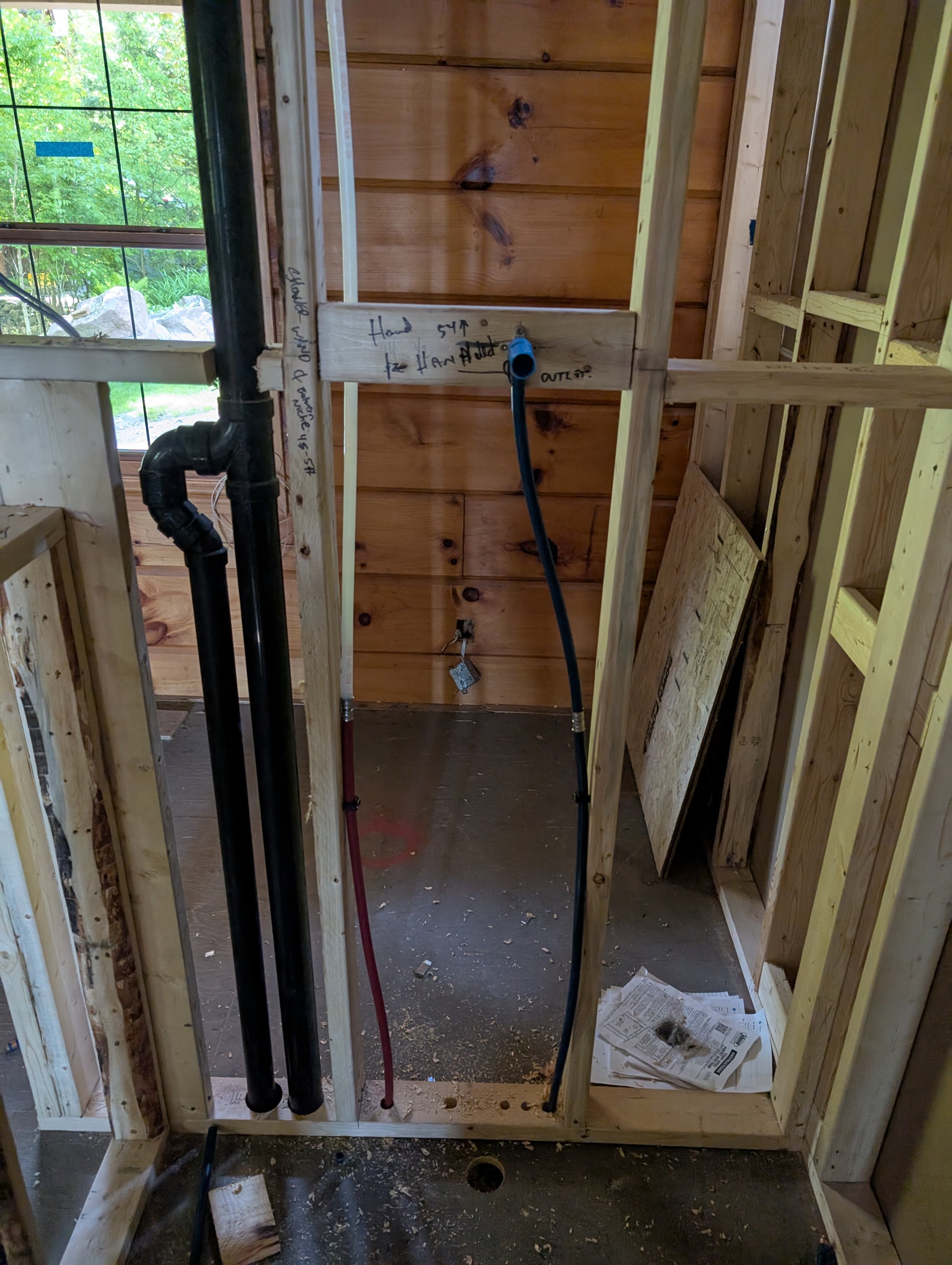 Interior of a house under construction with exposed wooden framing, plumbing pipes, a black drain pipe, a flexible water line, a window with green trees outside, and construction notes handwritten on the wood.