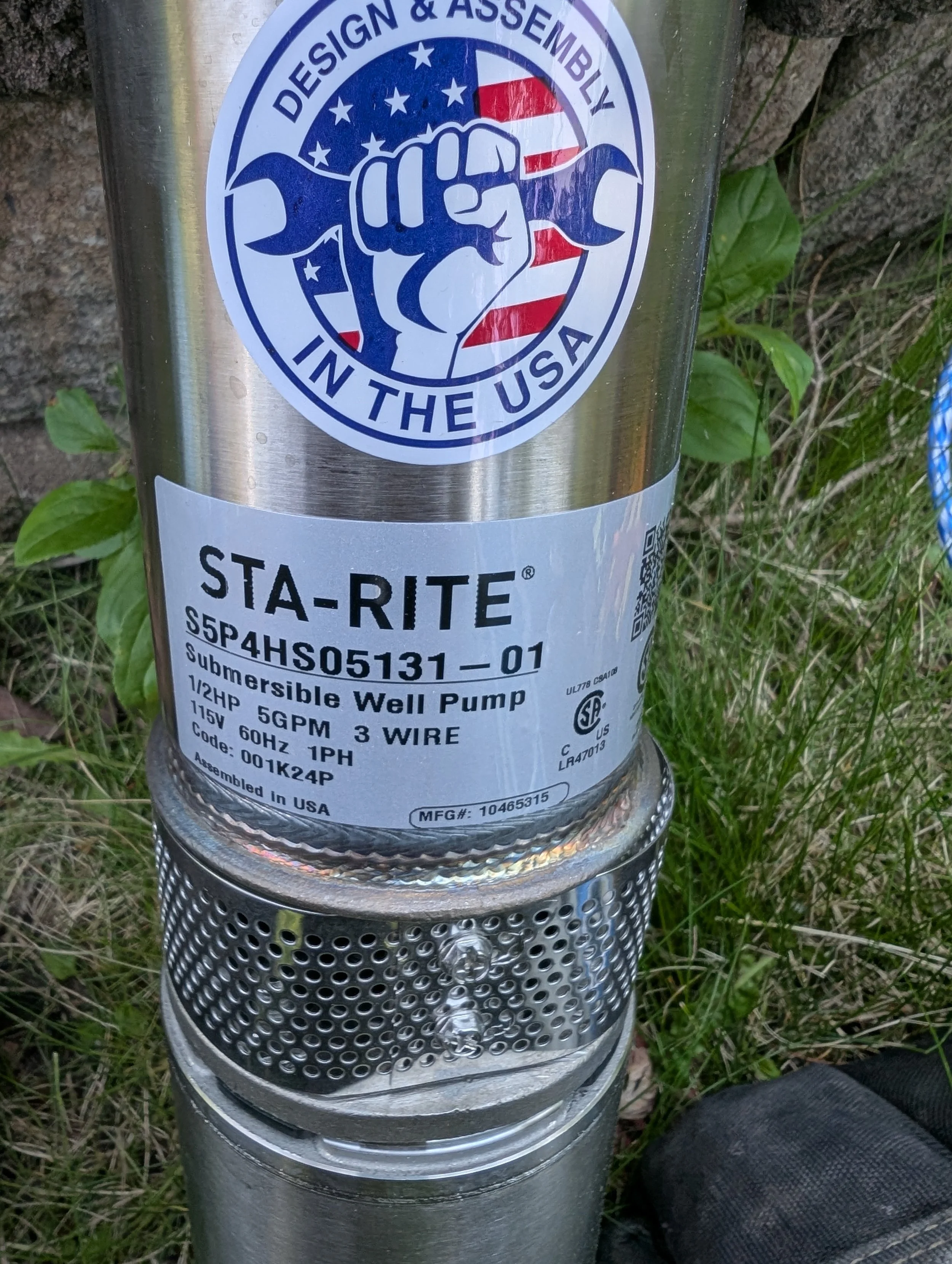 Close-up of a metal well pump with a patriotic sticker featuring a fist holding a wrench over a background of the American flag. The sticker reads 'Design & Assembly in the USA'.