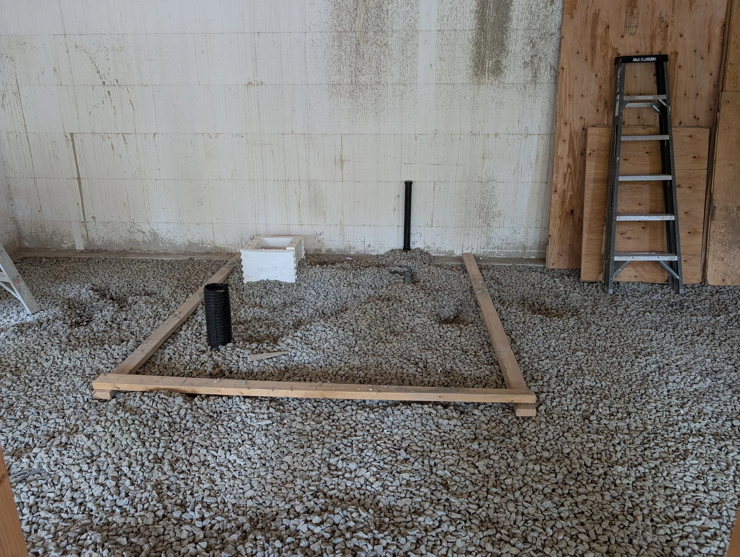 Construction site with gravel floor, partially built wooden frame, ladder, black pipe, and insulation on the wall.