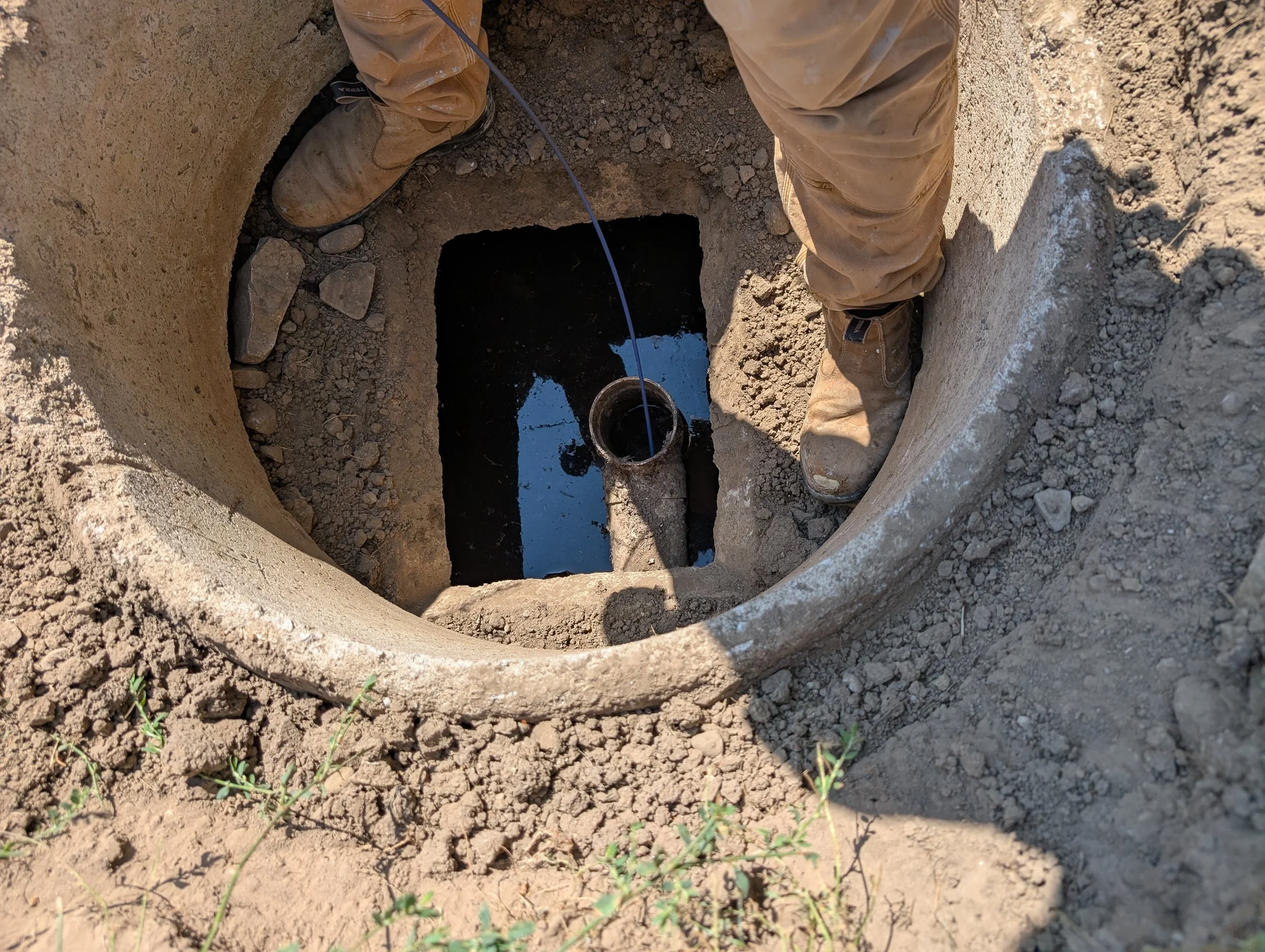A person in tan work boots and tan pants is working in a deep, square hole in the dirt, with some water collected at the bottom of the hole and pipes visible.