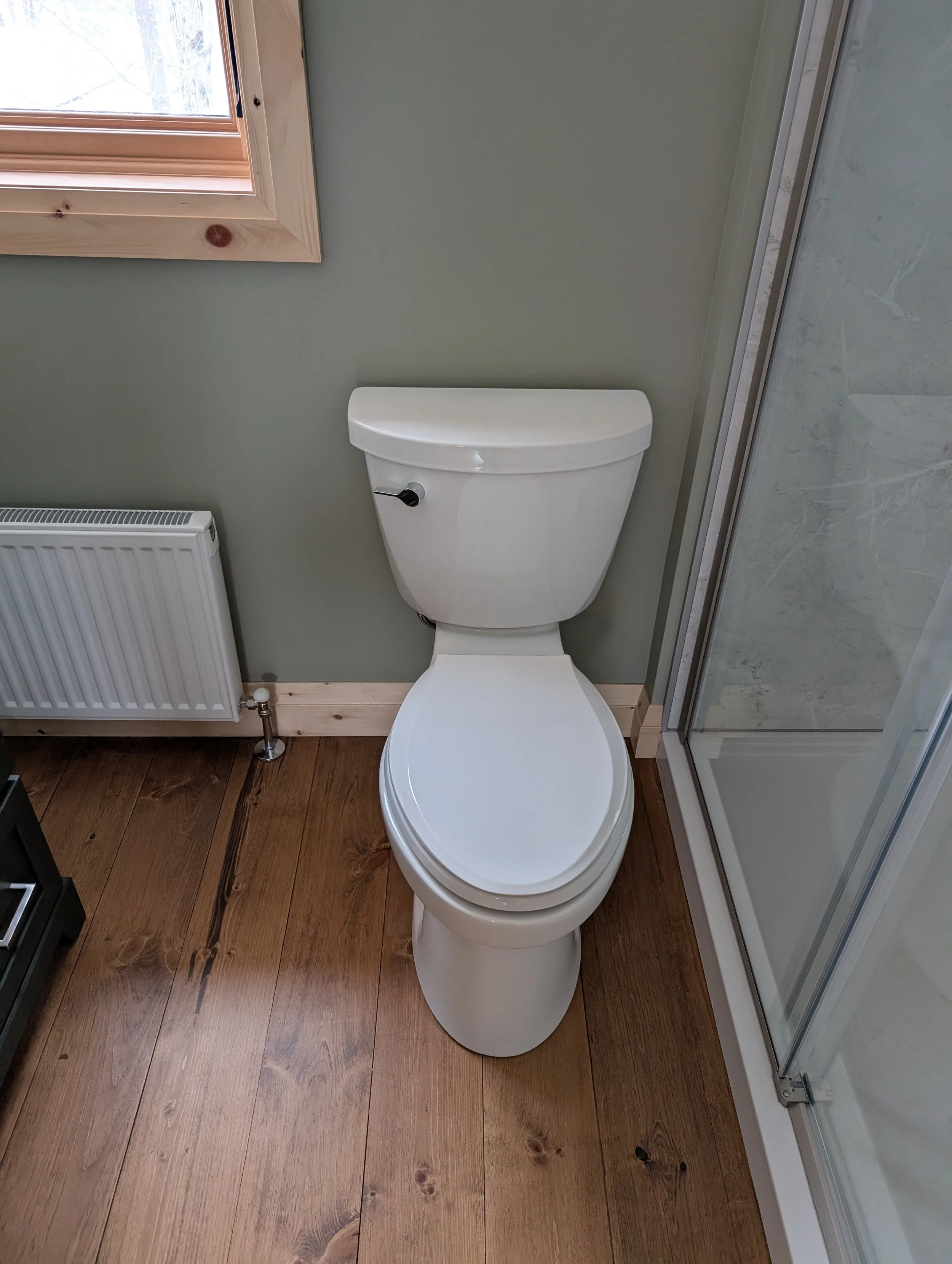 A bathroom with wooden flooring, a white toilet, a radiator, and a window with wooden trim.
