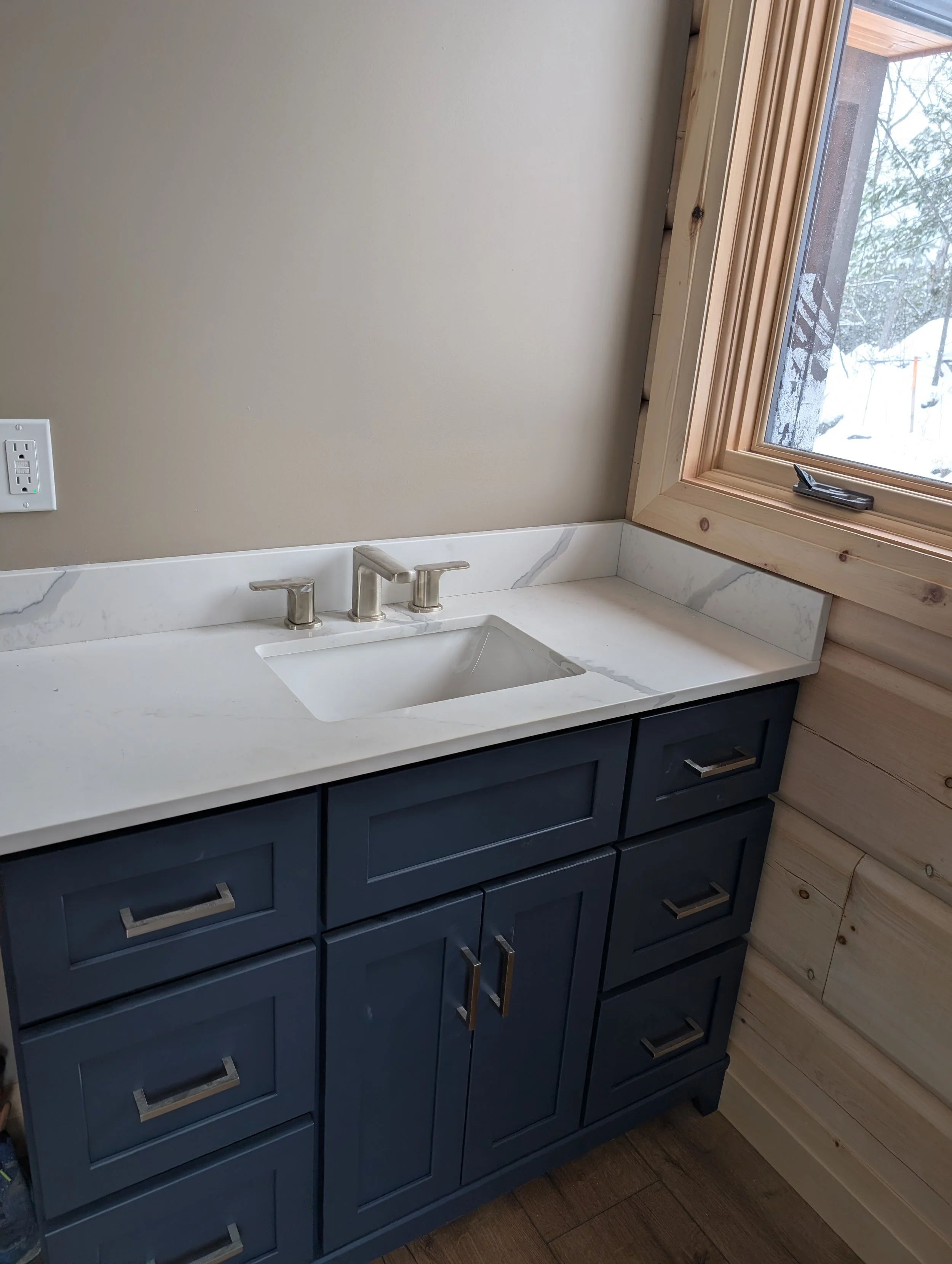 Close-up of a modern bathroom vanity with a white marble top, dark blue cabinets, and a gold-colored handle, situated below a wooden window with a snowy outdoor scene visible.