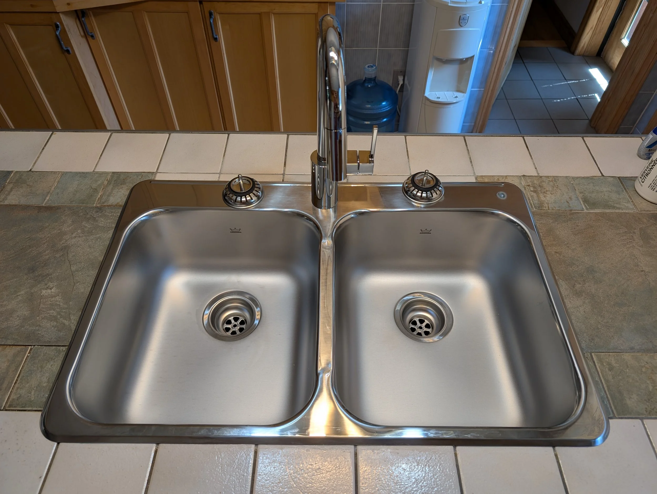 Kitchen sink with a chrome faucet, two basins, and beige and gray tiled countertops. In the background, a wooden cabinet, a water dispenser, and a doorway with a tiled floor are visible.