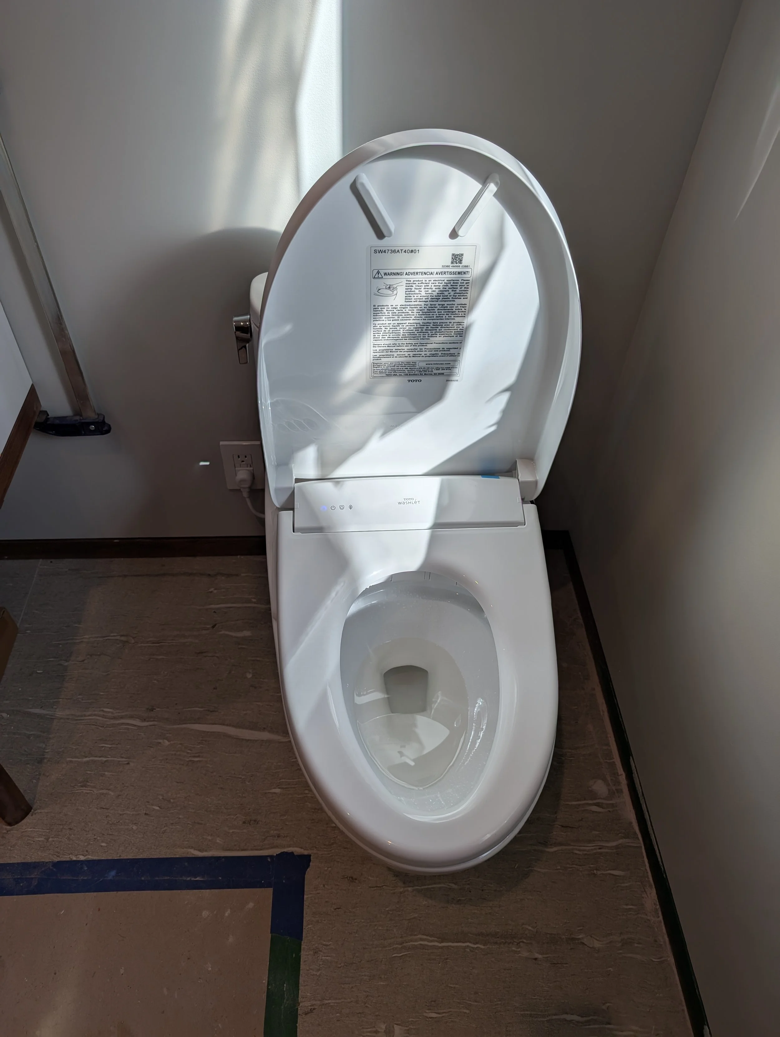 A modern white toilet with an open lid in a bathroom with wood-look flooring. There is a wall outlet and a power cord nearby, and a sticker with warnings on the lid.