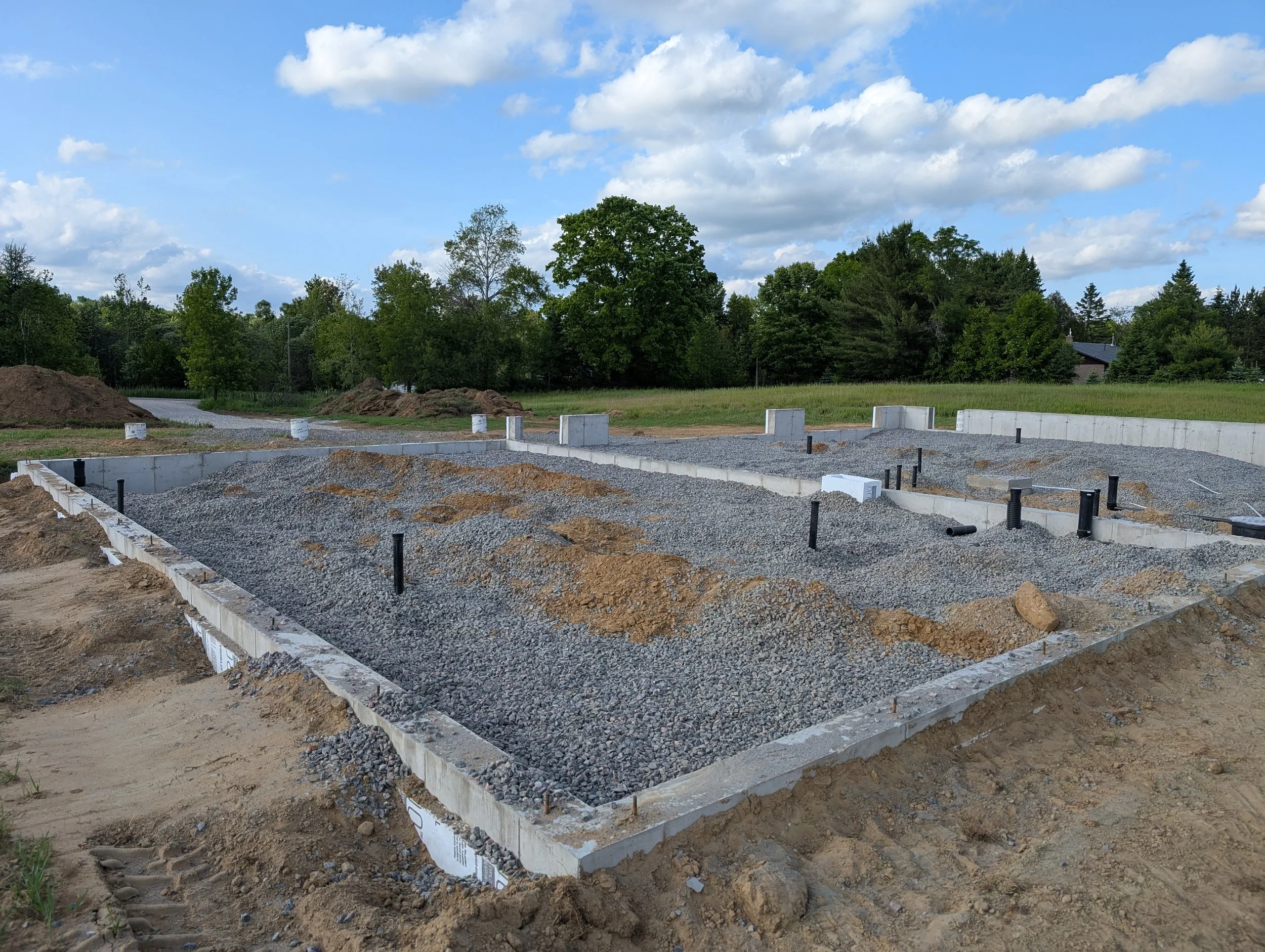 A construction site with foundation and gravel, surrounded by dirt, with a backdrop of trees and a blue sky with clouds.
