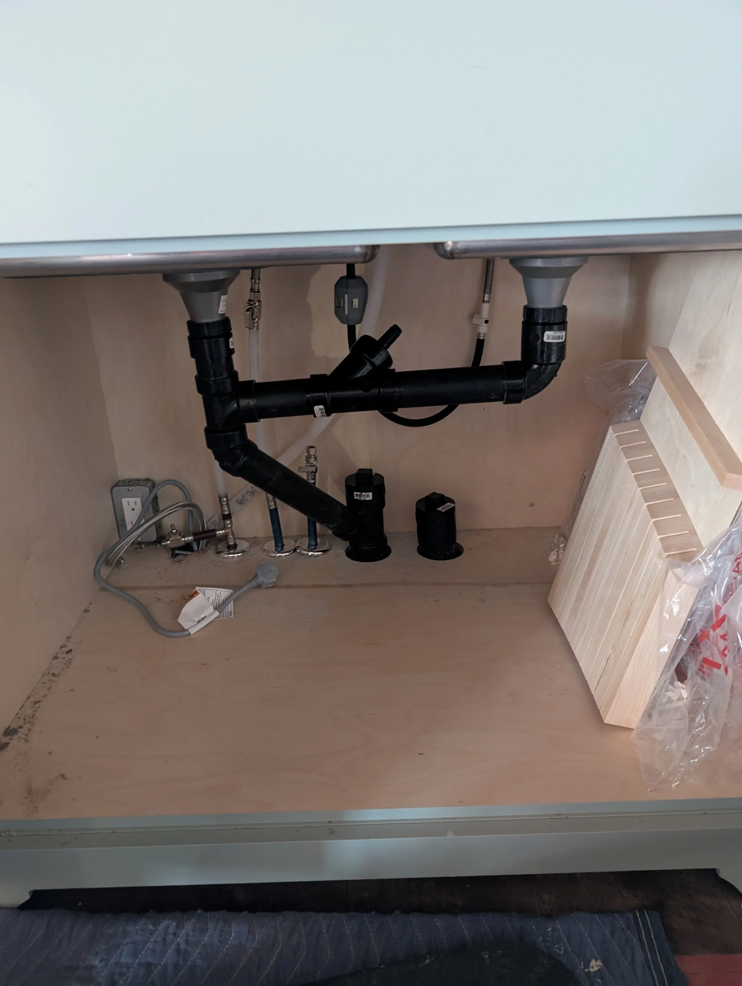 Underneath a kitchen sink, plumbing pipes including a black P-trap, a white drain pipe, and various fittings are visible. There are two electrical outlets on the wall near the floor, and a section of wooden countertop with a backsplash is shown on th