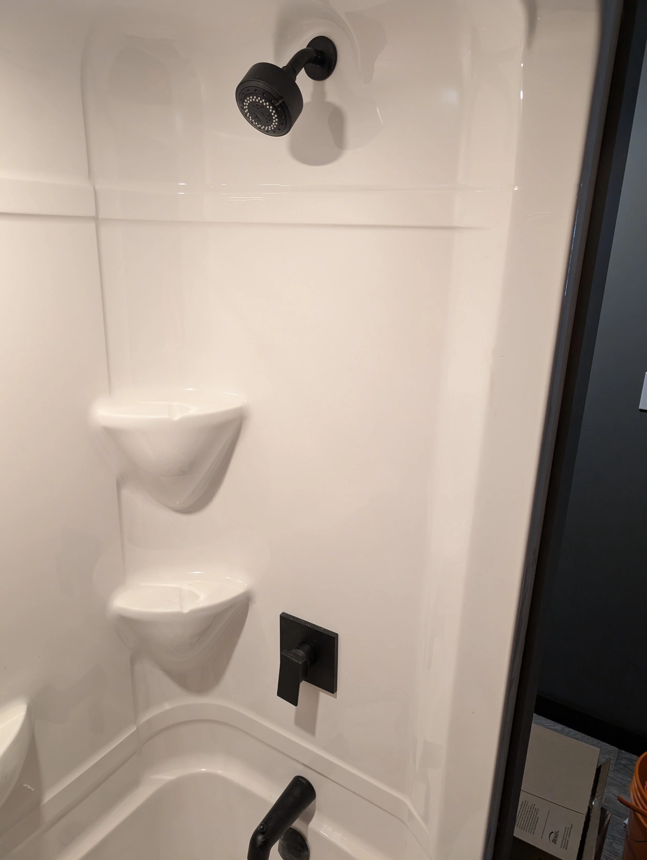 A black showerhead installed on a white shower wall with built-in shelves and a black faucet handle below.