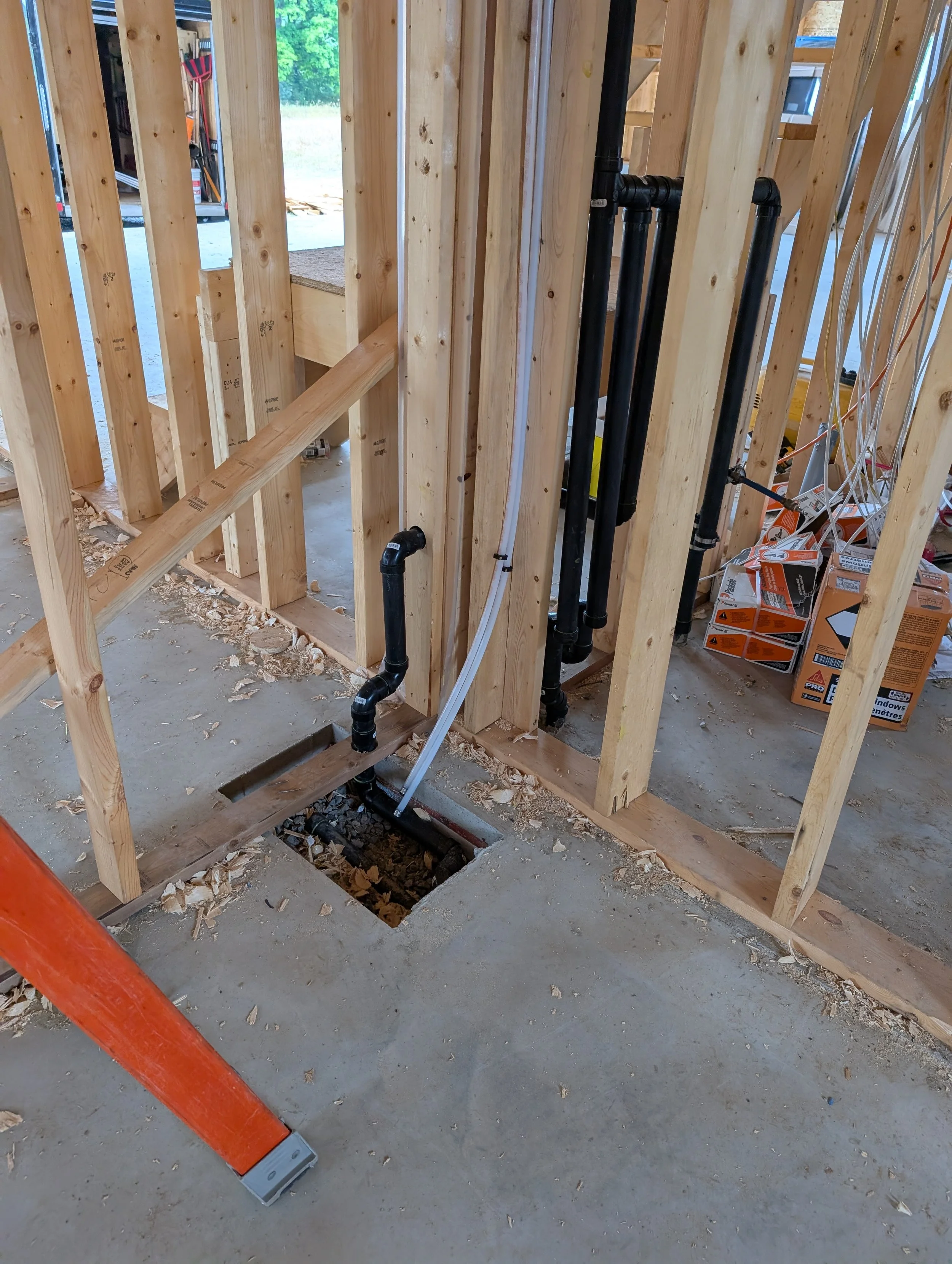 Interior view of a house under construction with exposed wooden framework, plumbing pipes, electrical wiring, and a rectangular hole in the concrete floor for plumbing installation.