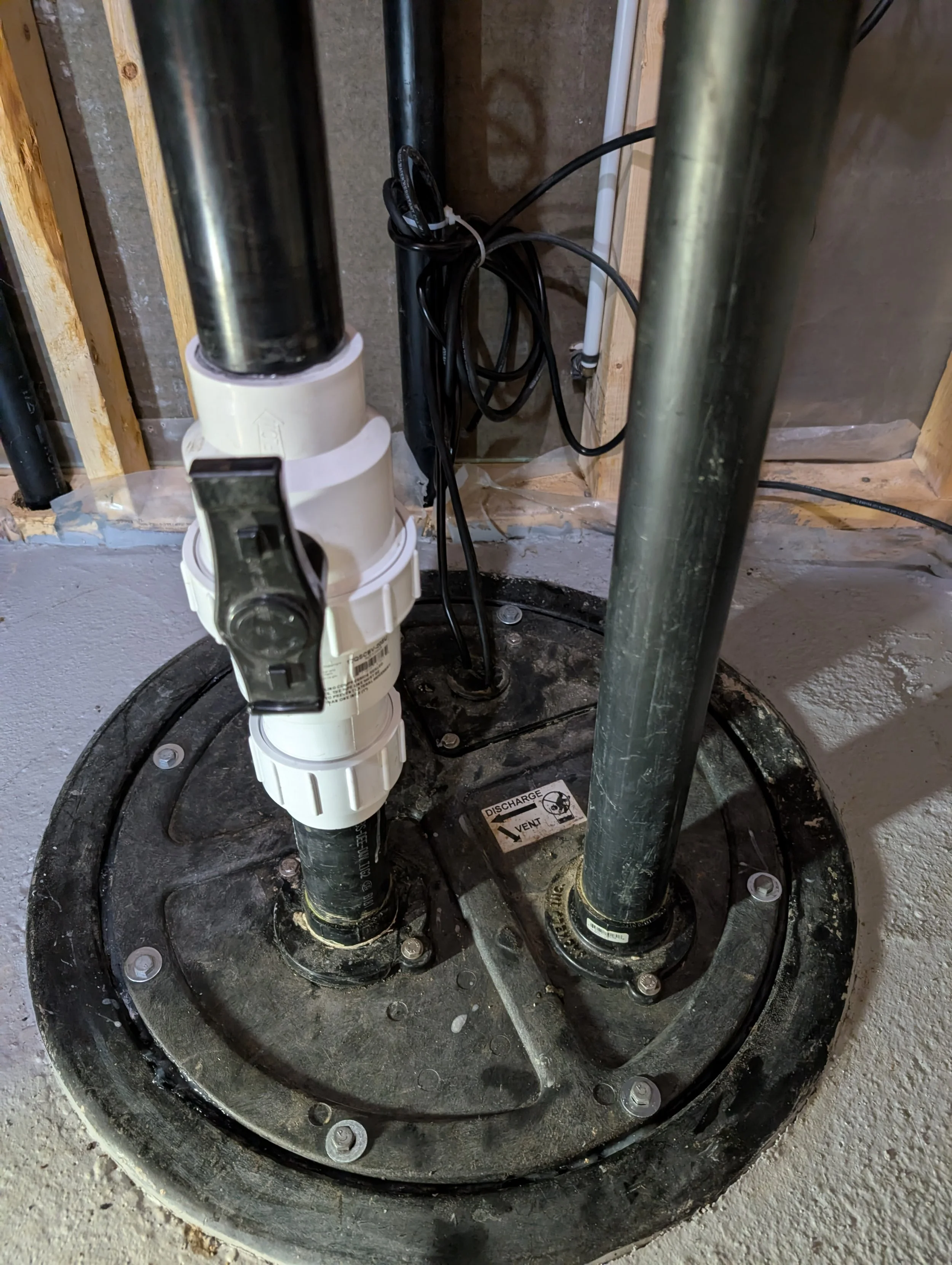 Close-up of a plumbing setup in a construction or repair site, featuring pipes, electrical wires, and a large black circular base with bolts and a warning label.