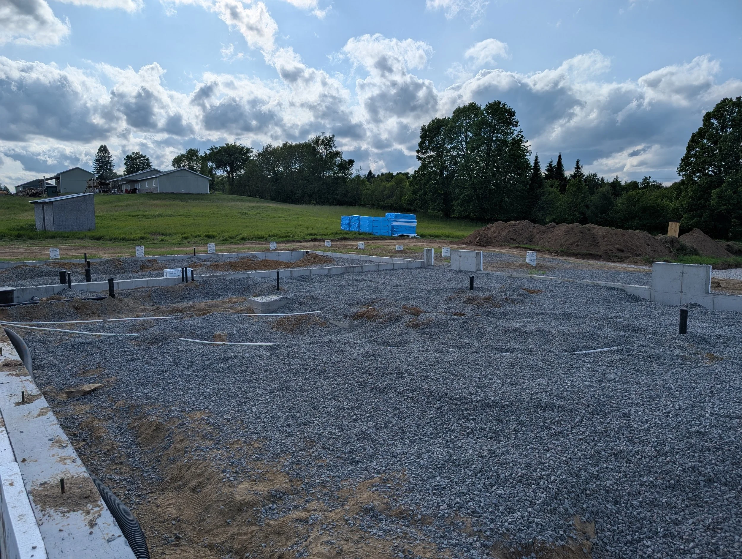 Construction site with gravel and concrete foundation, pipes, and construction signs, with a grassy hill, trees, and houses in the background under a partly cloudy sky.