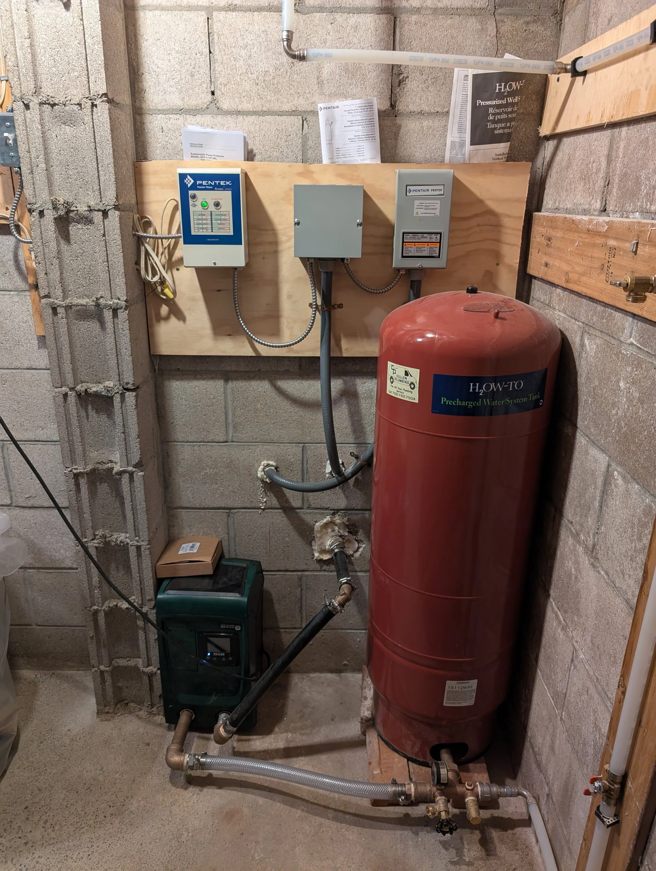 An indoor utility room with various plumbing and water system components, including a red water pressure tank, electrical control panels, and pipes running along the wall.