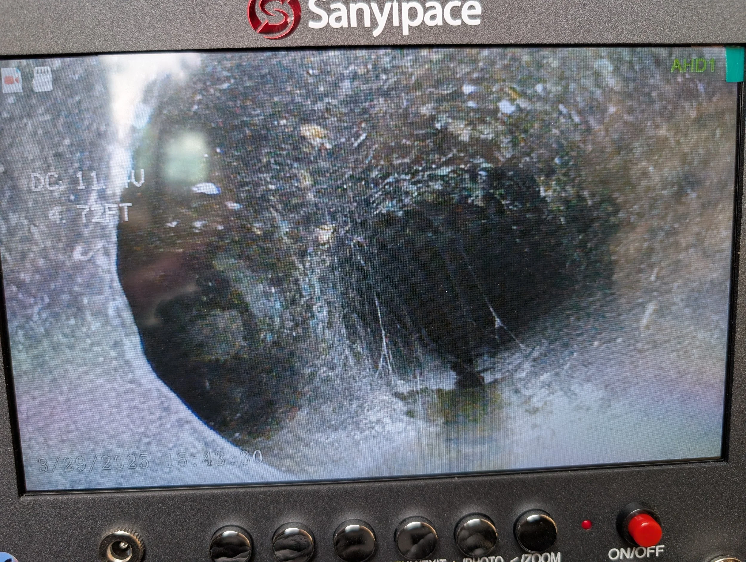 Close-up of a Sanypace device screen showing a blurred outdoor soil and plant scene. The screen displays the date and time, December 11th at 3:43:30 PM.