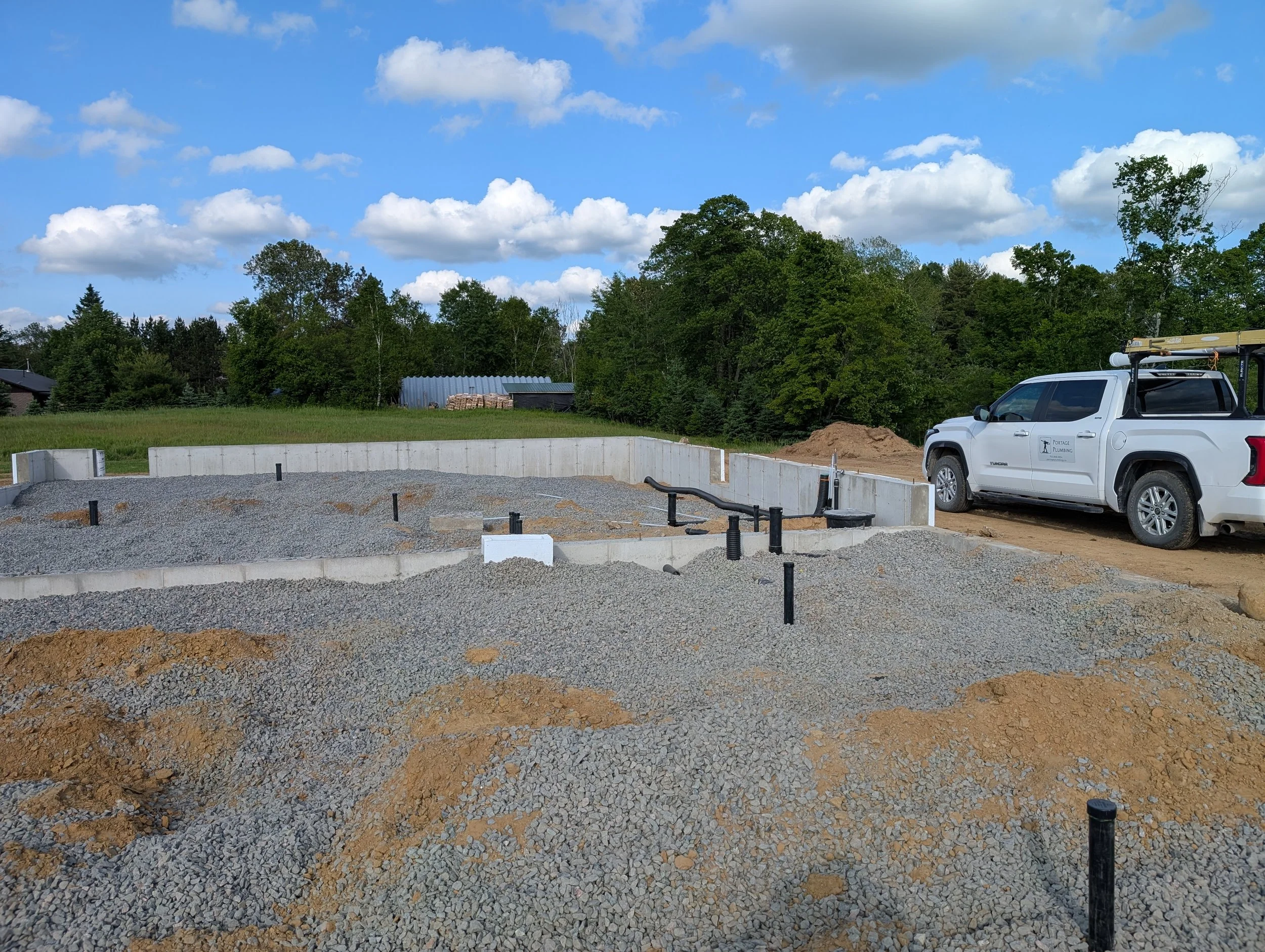 Construction site with gravel and dirt, concrete foundation, a white pickup truck with a ladder rack, trees and cloudy sky in the background.