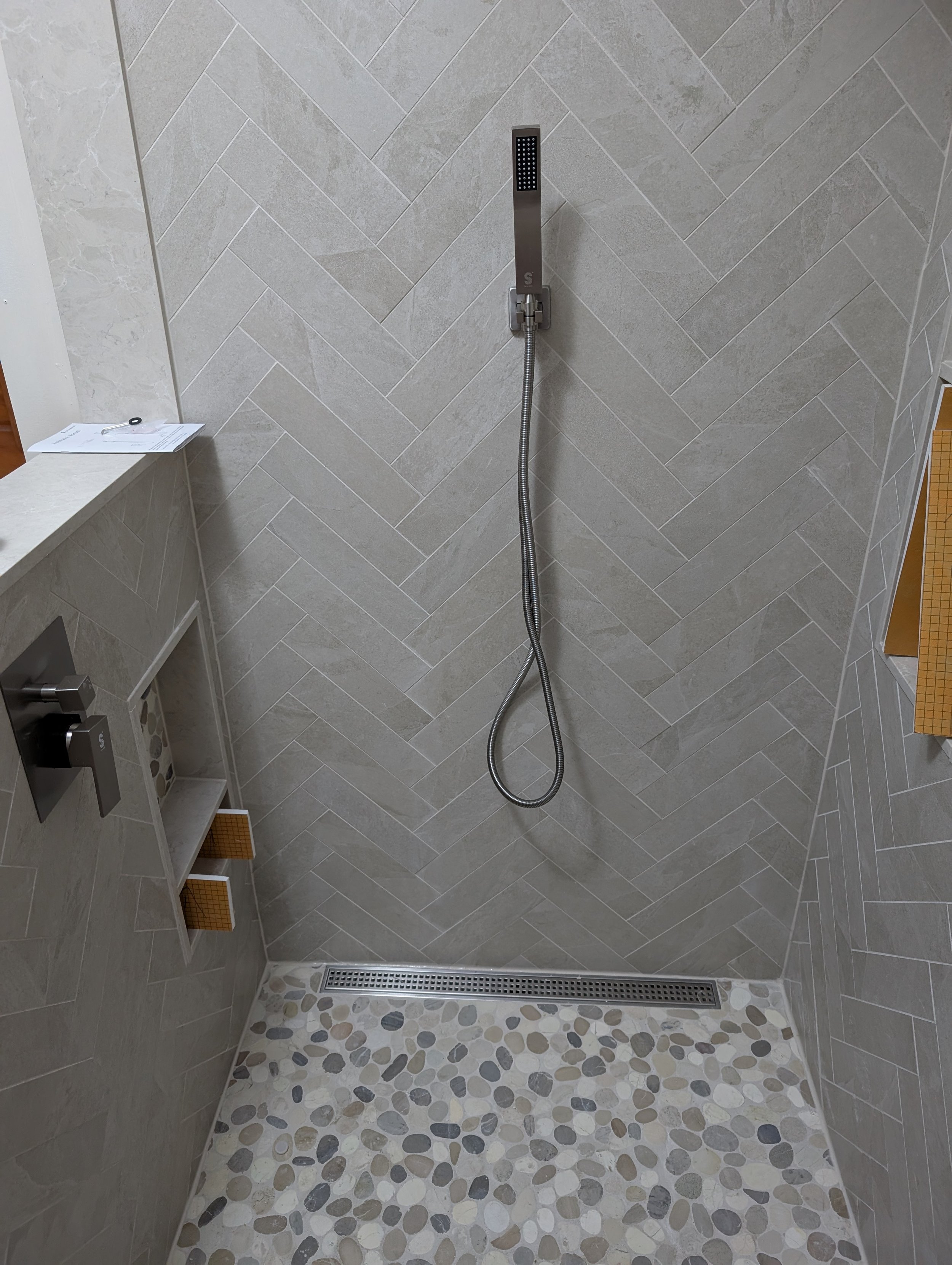 View of a modern walk-in shower with grey tiles arranged in a herringbone pattern on the walls, a handheld showerhead, a small corner shelf with toiletries, and a pebble tile floor with a linear drain.