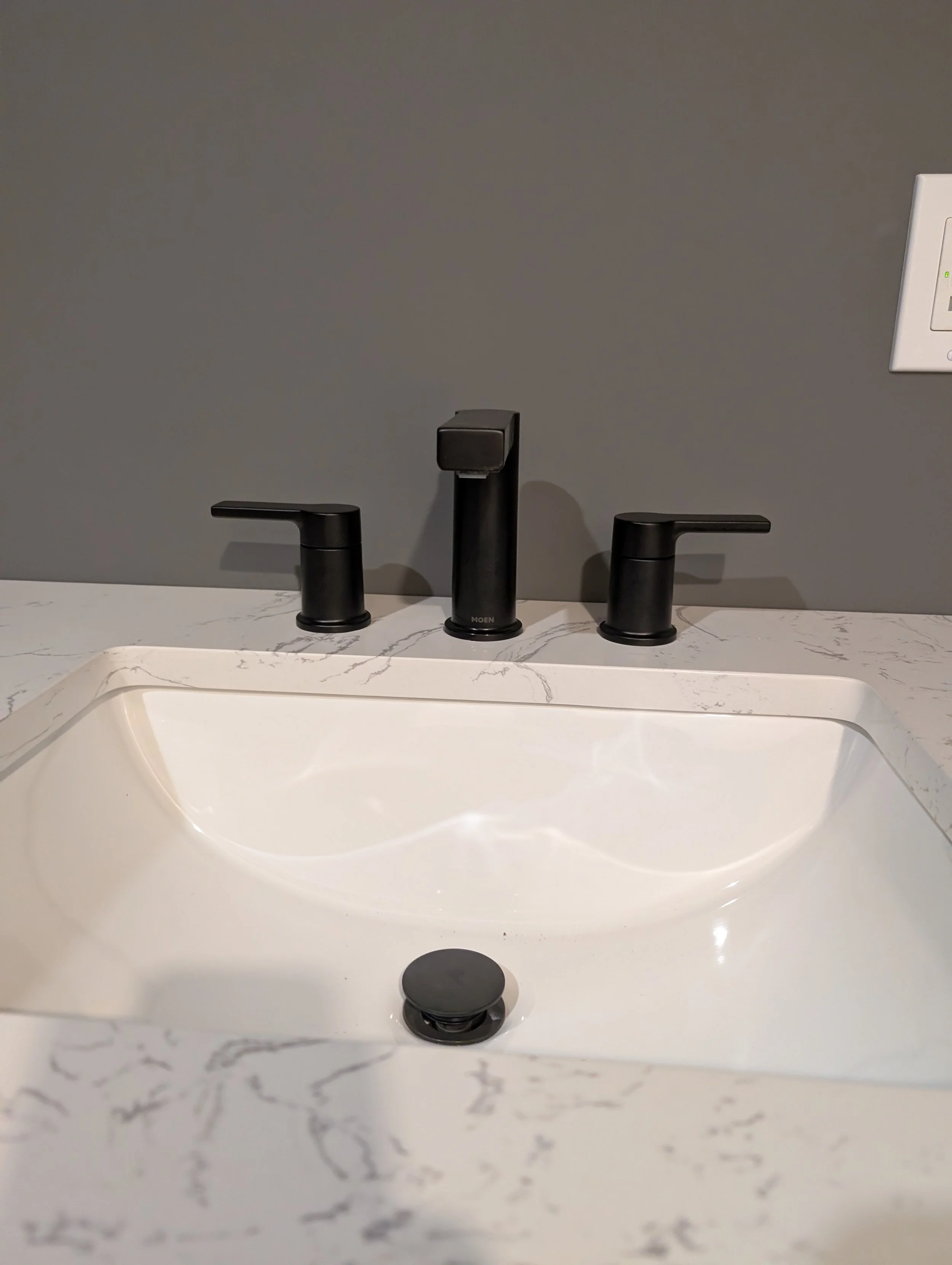 A modern black bathroom faucet set with two handles above a white marble countertop and a rectangular undermount sink against a gray wall with a white switch plate.