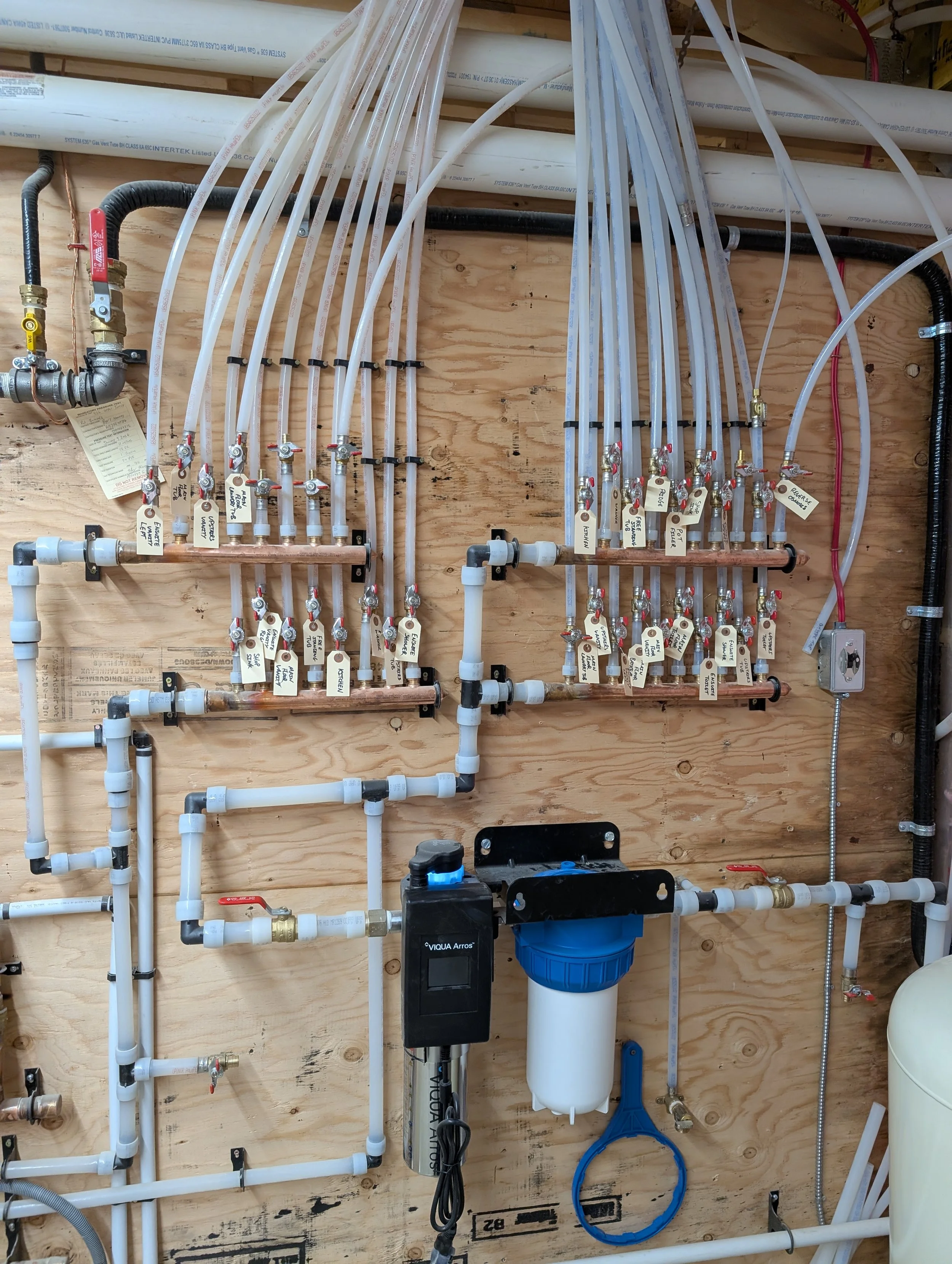 Water filtration and plumbing system with various white PVC pipes, valves, and a blue water filter attached to a wooden wall.