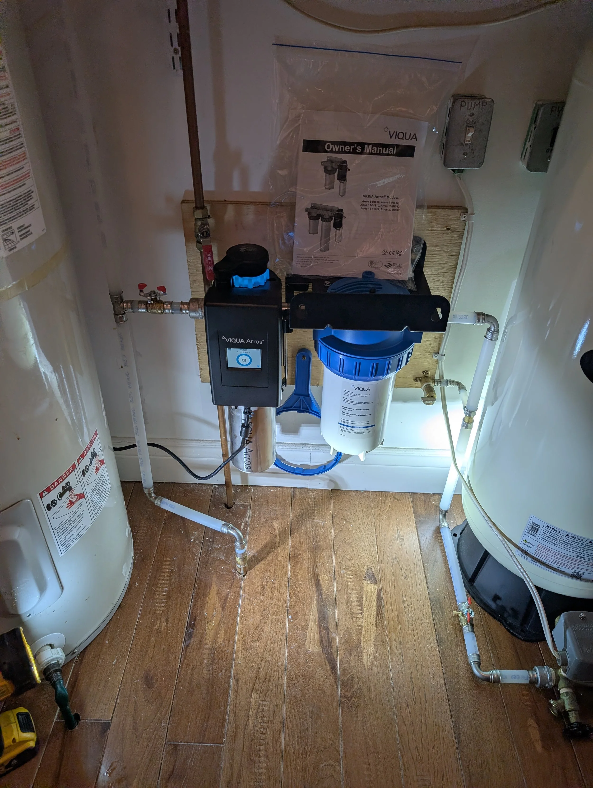 Image shows a plumbing setup with a water filter system, pipes, a water pump, and a manual. There is a tank on the left and another on the right, along with a wooden panel on the wall.