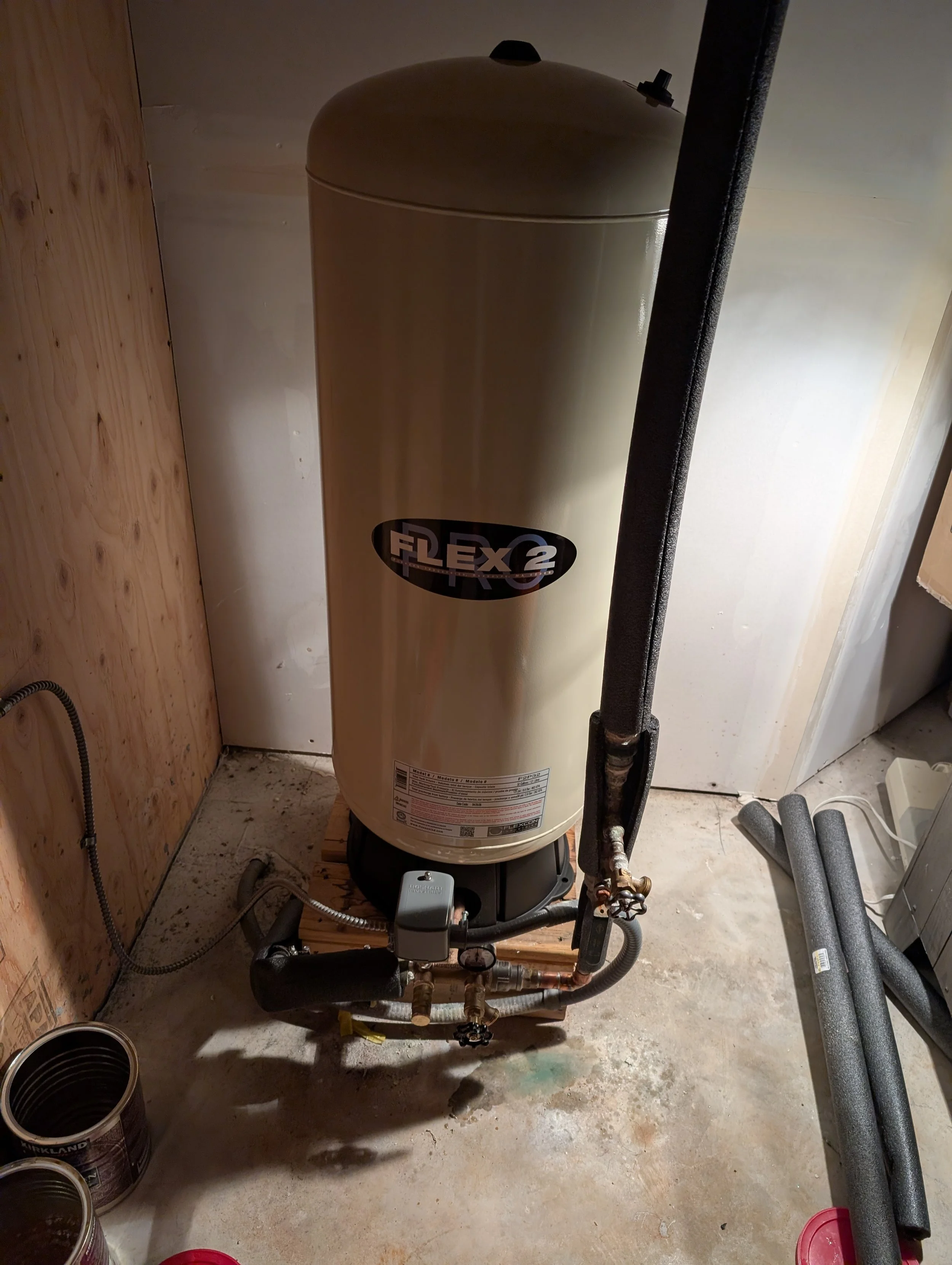 A water heater labeled Flex 2 installed in a basement with wooden and drywall walls, connected to various pipes and a valve. Multiple cans of paint are on the floor nearby.