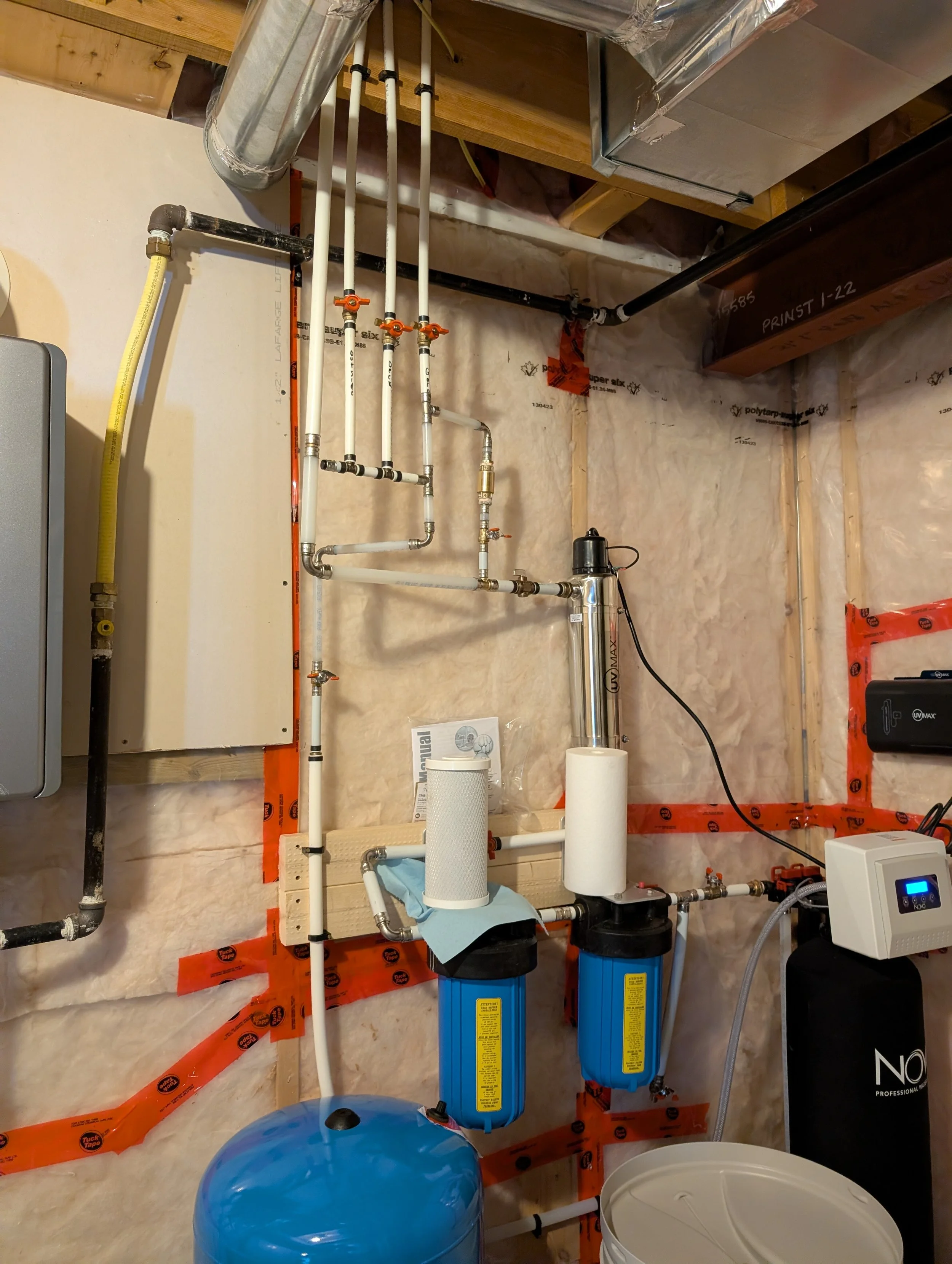 Piping and equipment setup in a basement utility room, including water filtration system with two blue filters, a black pump, white PVC pipes with valves, and other plumbing components.