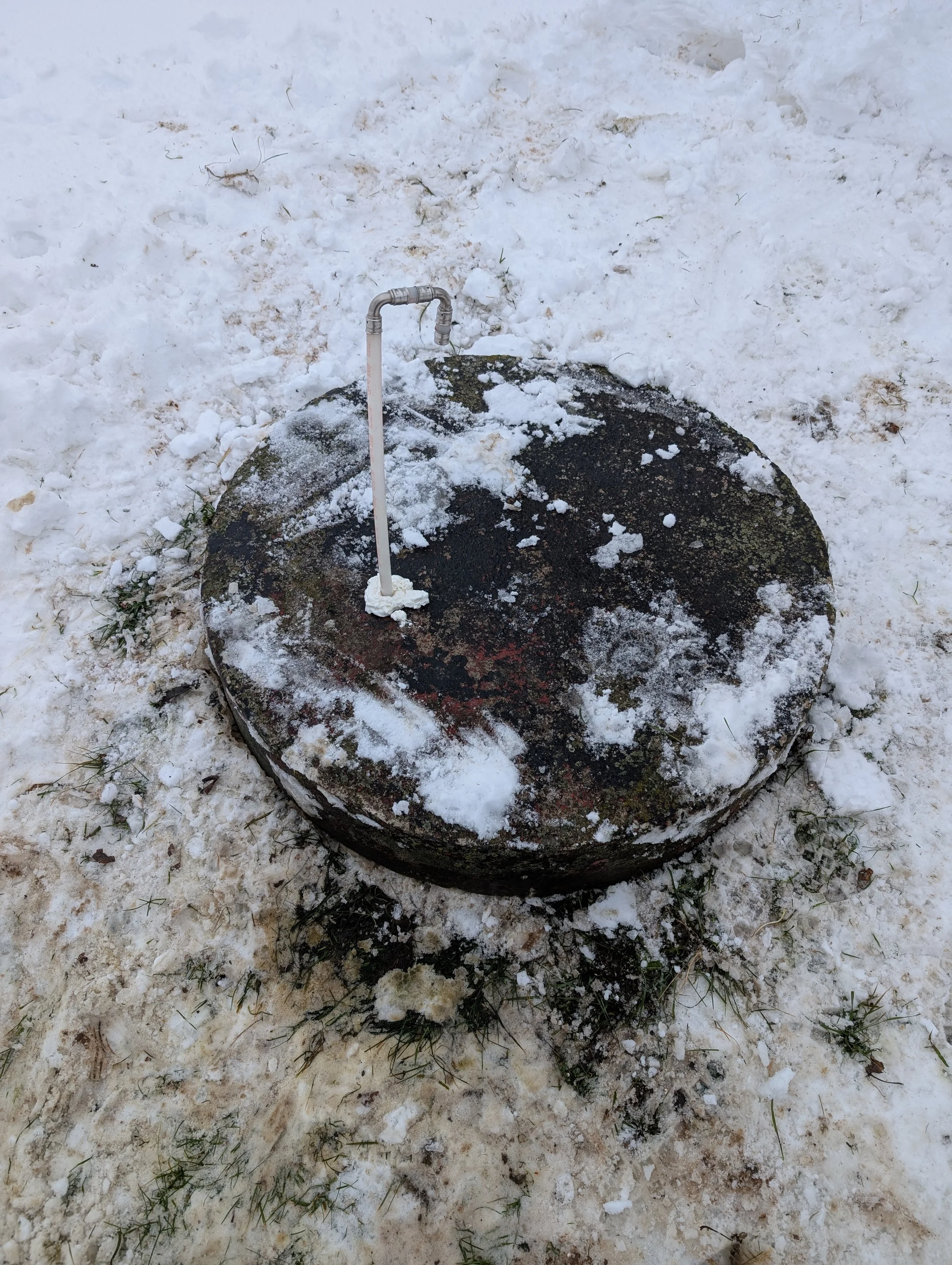 A snow-covered ground with a large, black, mossy rock and a metal outdoor tap attached to a pipe on top of the rock.