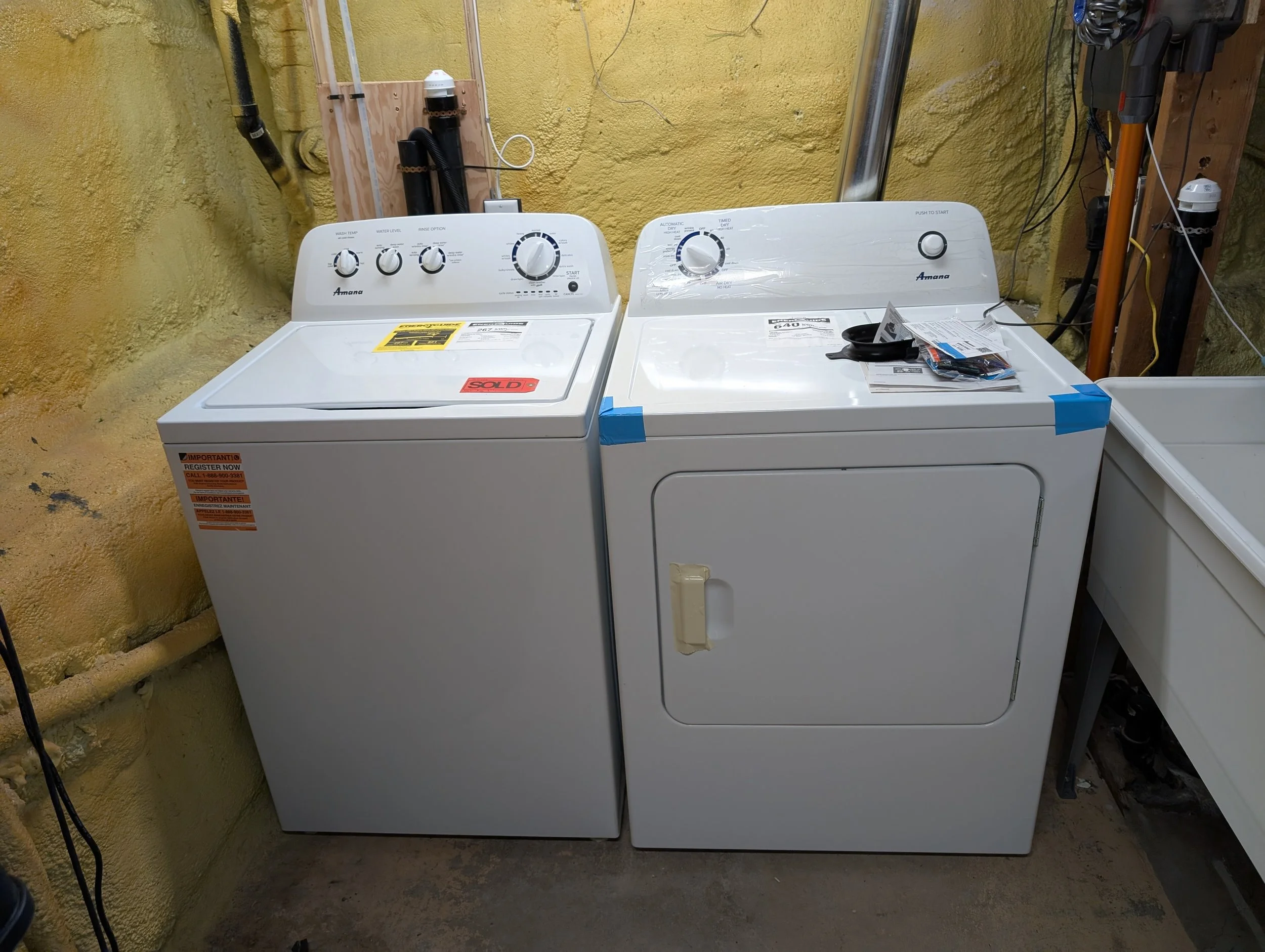 A laundry room with a white washing machine and dryer next to each other against a yellow textured wall. Various black and yellow pipes and wires are visible above the appliances, and there is a pair of black scissors and some paper on top of the dry