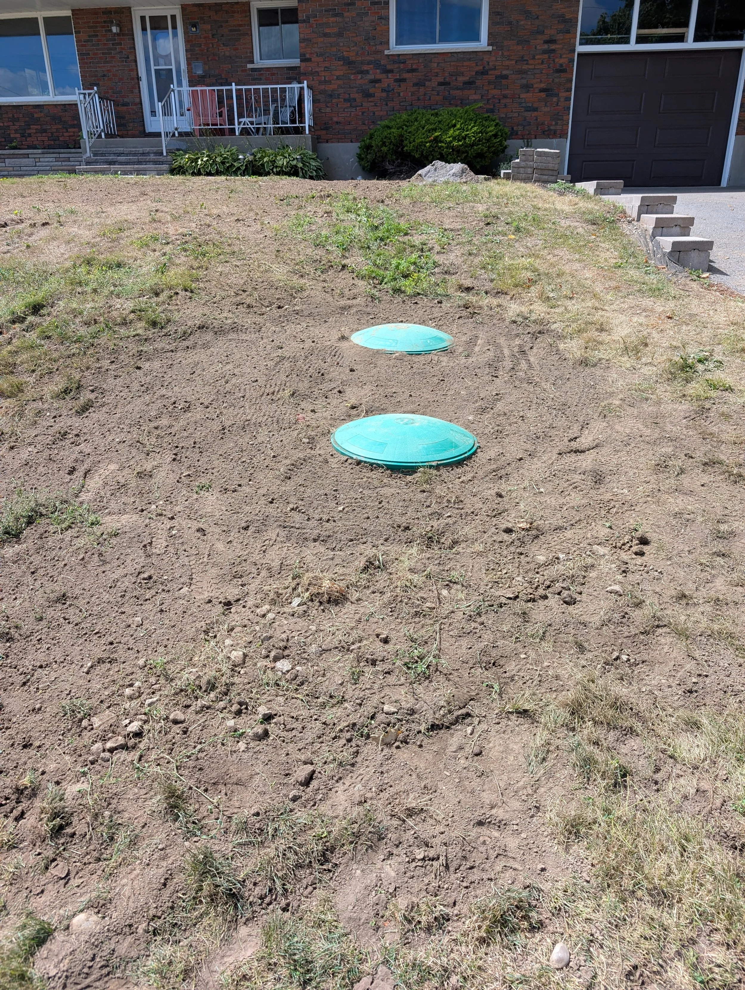 A front yard with dirt and patches of grass in front of a brick house. There are concrete steps leading up to a glass door with a small porch. Two round blue covers are embedded in the dirt, likely for utility access.
