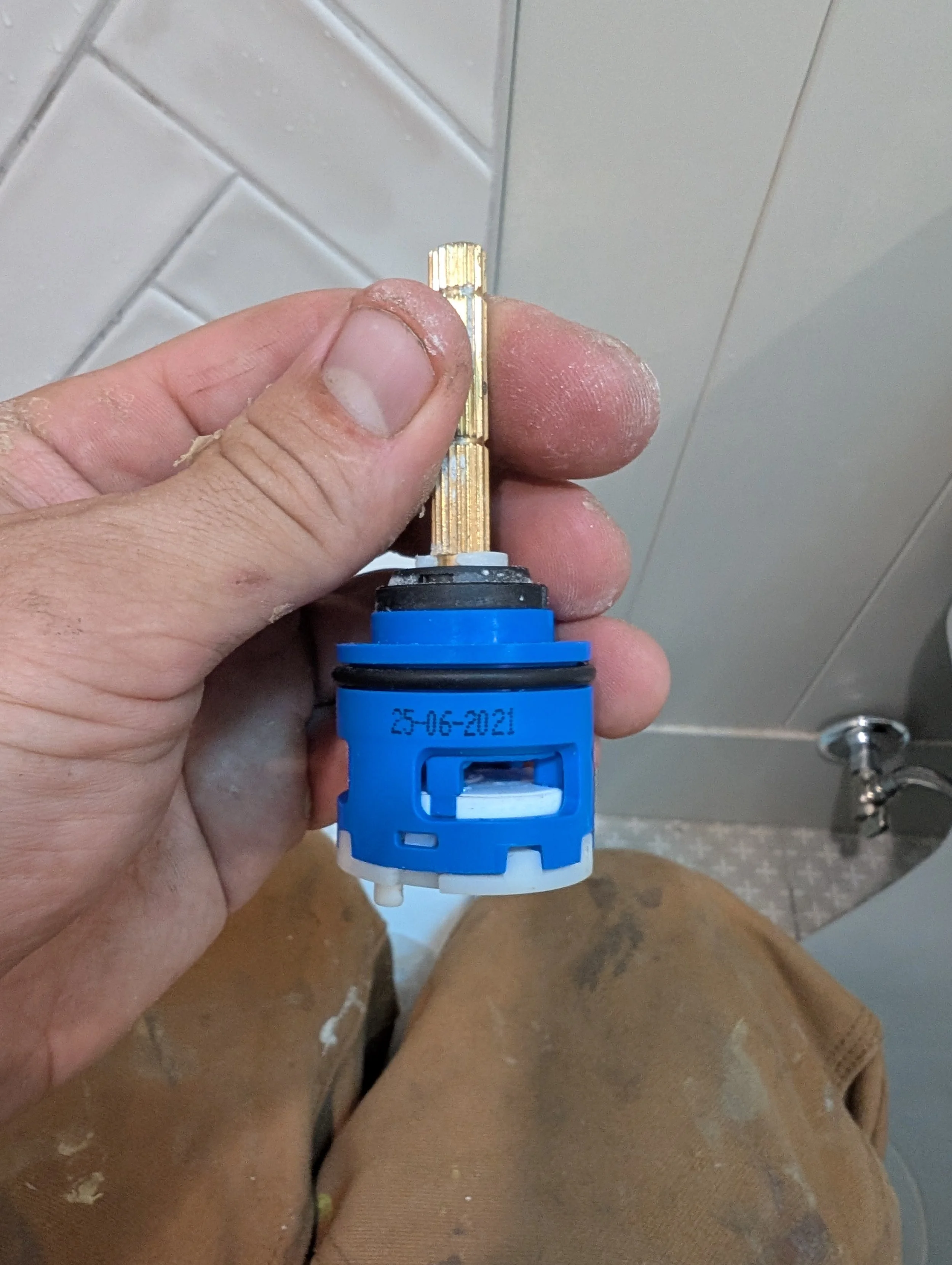 Close-up of a hand holding a small metallic component over a person's legs and brown tiled floor, with a bathroom sink faucet in the background. The component has a date stamped on it: 25-06-2021.
